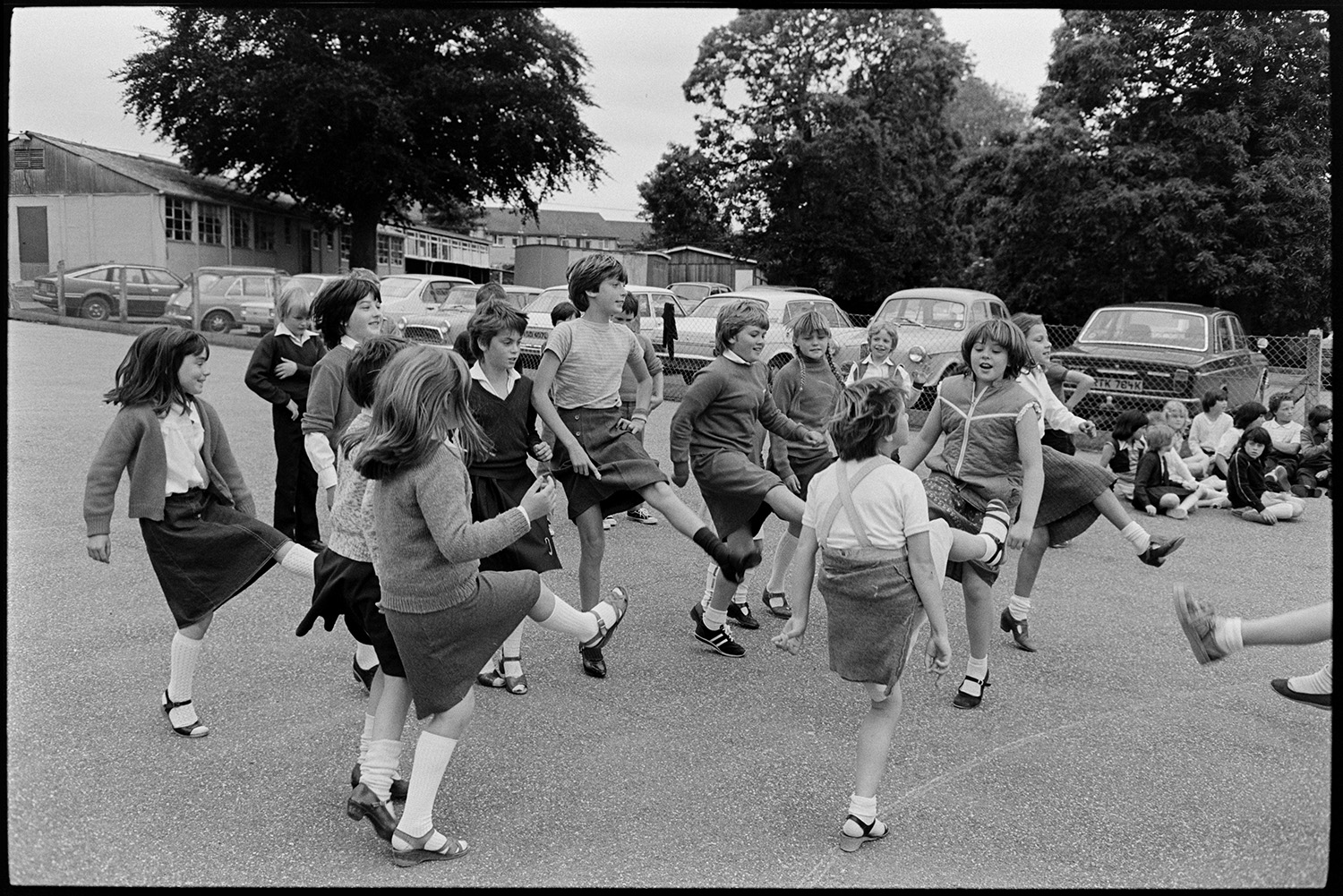 Schoolchildren singing, dancing with folk duo. 
[Schoolchildren dancing in a playground in Barnstaple, at a folk workshop run by the folk duo Sam Richards and Tish Richards. Cars are parked behind the playground and school buildings can be seen in the background.]