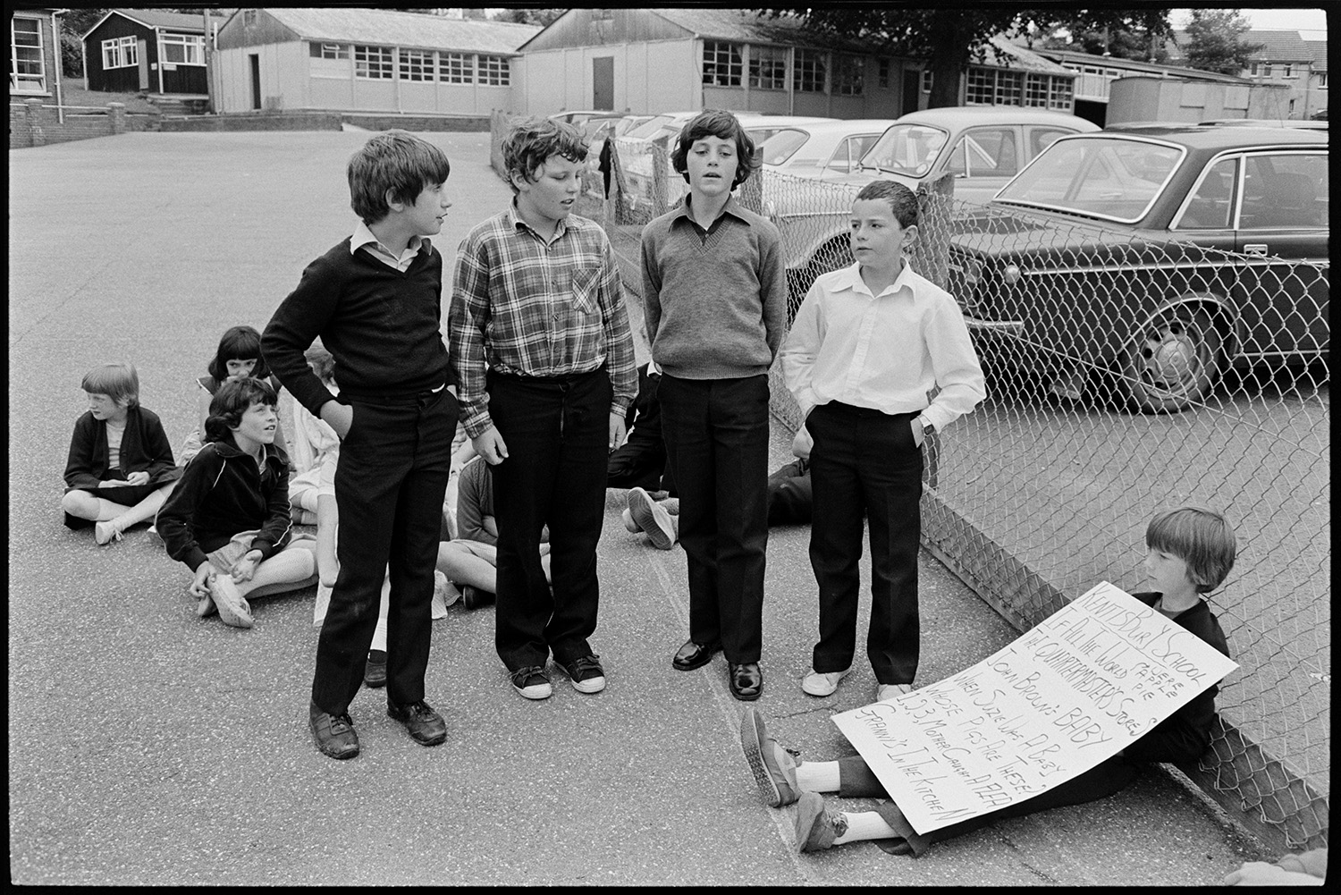 Schoolchildren singing, dancing with folk duo. 
[Four boys singing in a folk workshop, run by folk duo Sam Richards and Tish Richards, in the playground of a school in Barnstaple. One boy is holding a poster, possibly with song words. Other children are sat down listening. Parked cars and school buildings can be seen in the background.]