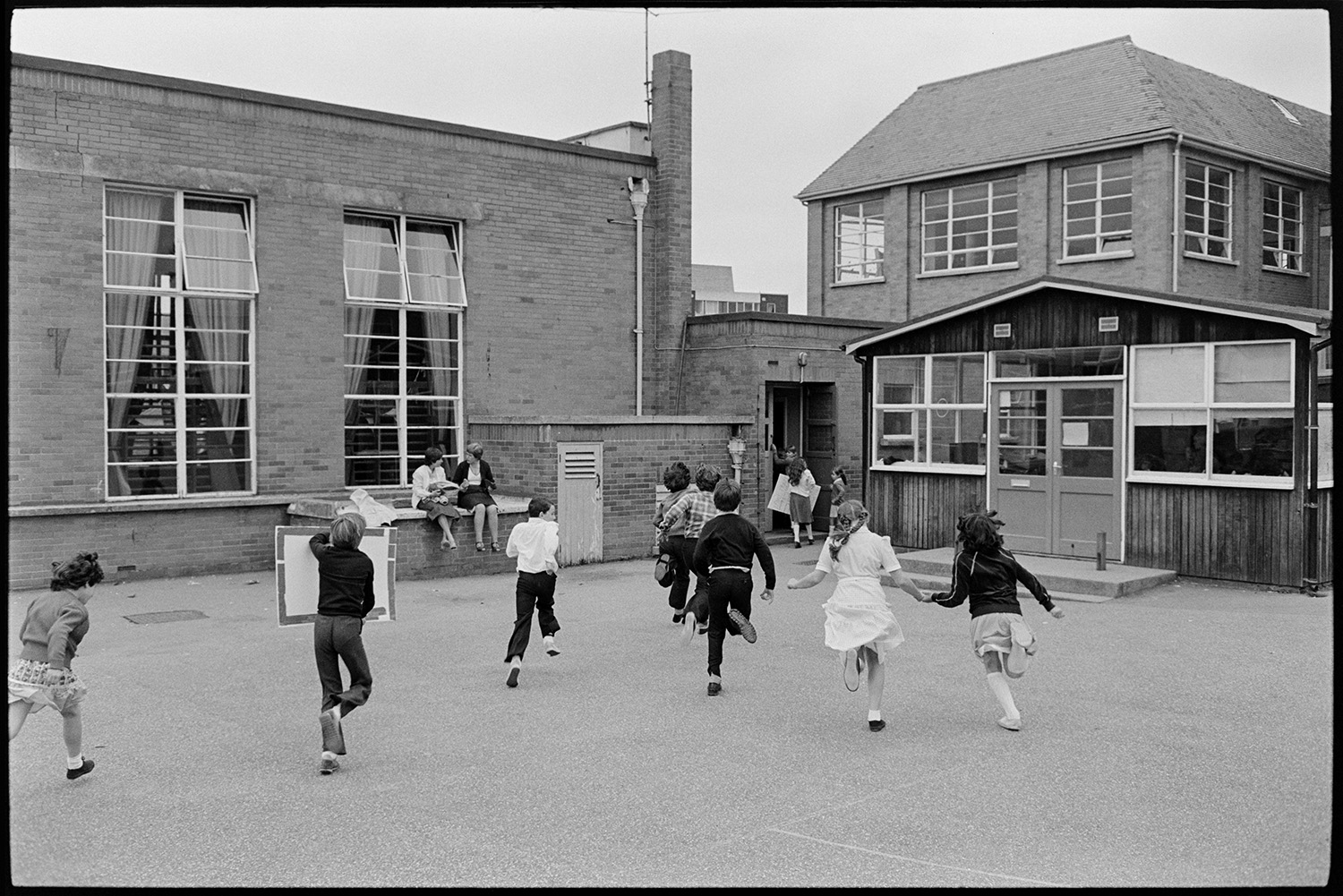 Schoolchildren singing, dancing with folk duo. 
[Children running across a playground towards a school building, at a school in Barnstaple. Possibly after taking part in a folk workshop run by folk duo Sam Richards and Tish Richards.]