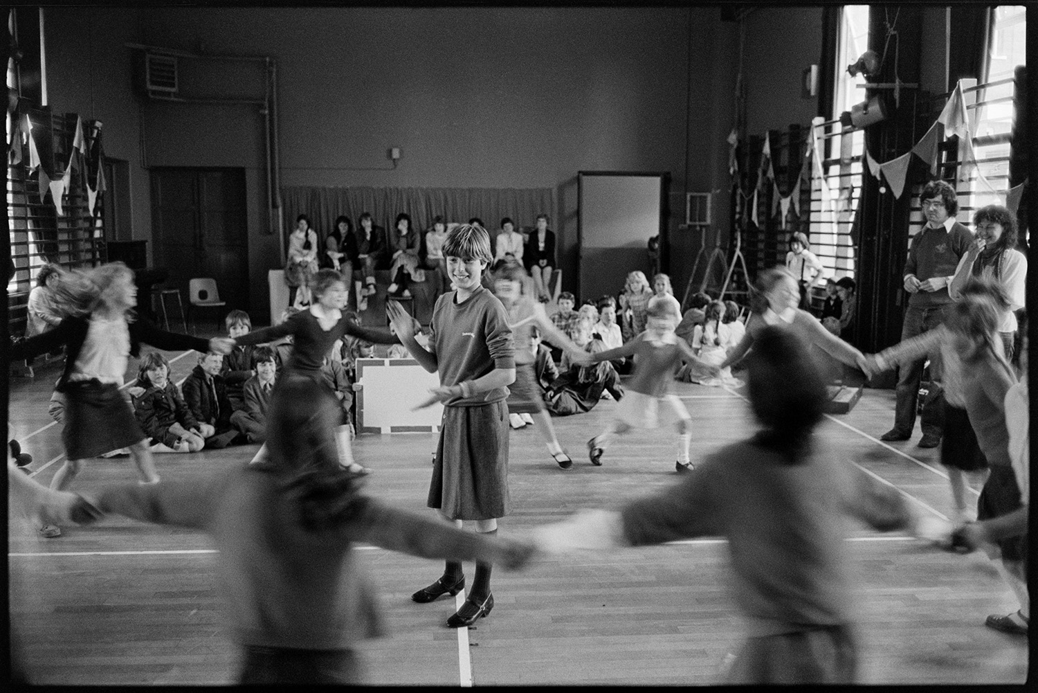 Schoolchildren singing, dancing with folk duo. 
[Children dancing in a circle with one child clapping in the middle, at a folk workshop in a school hall in Barnstaple, run by folk duo Sam Richards and Tish Richards. Other children are sat watching in the background.]