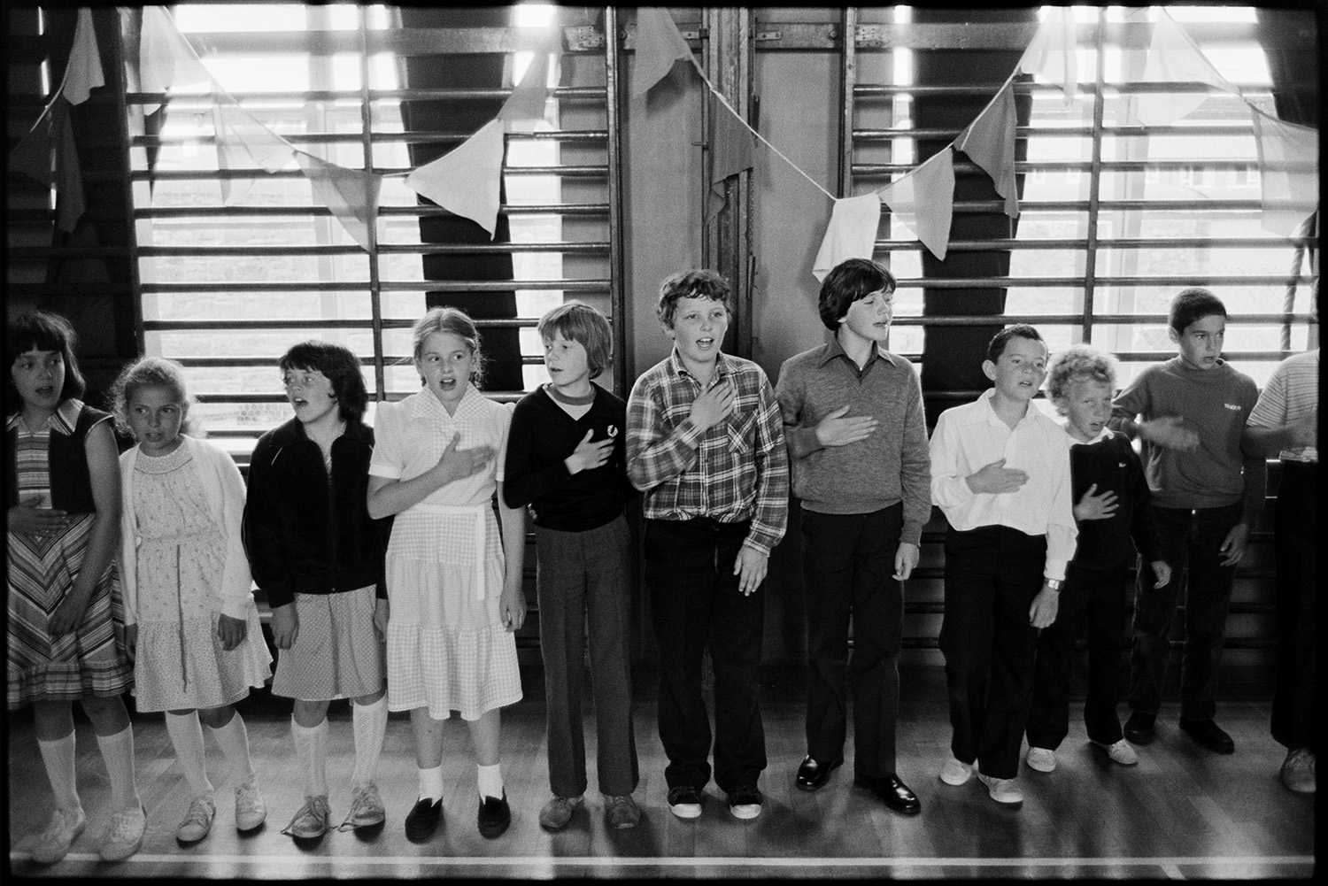 Schoolchildren singing, dancing with folk duo. 
[Children singing with their hands across their hearts in a folk workshop in a school hall in Barnstaple, run by folk duo Sam Richards and Tish Richards. The gymnastic equipment behind them is decorated with bunting.]