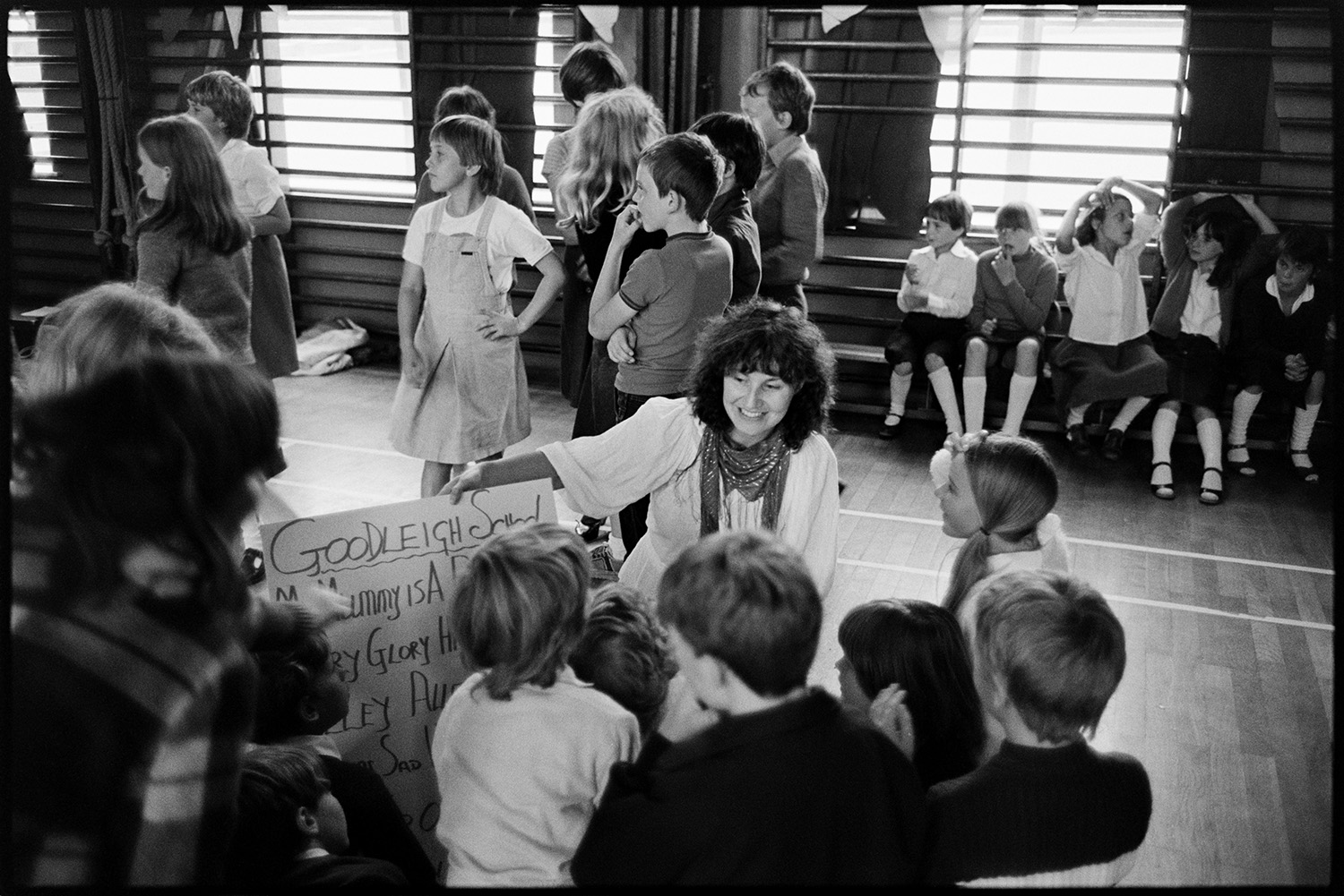 Schoolchildren singing, dancing with folk duo. 
[Children leaning folk songs at a folk workshop in a school hall in Barnstaple being run by folk duo Sam Richards and Tish Richards. Tish Richards is holding the words of a song up for a group of children.]