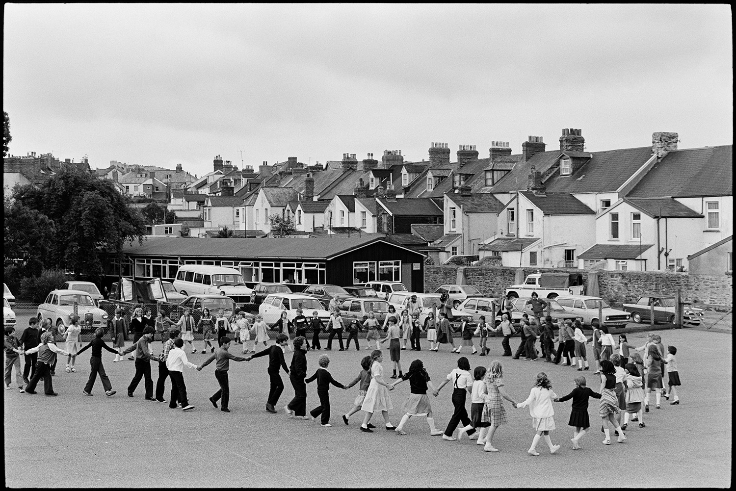 Schoolchildren dancing in playground  with folk duo. 
[Children dancing in a large circle in a folk workshop, run by Sam Richards and Tish Richards, in a school playground in Barnstaple. They are being filmed. Terraced houses can be seen in the background.]