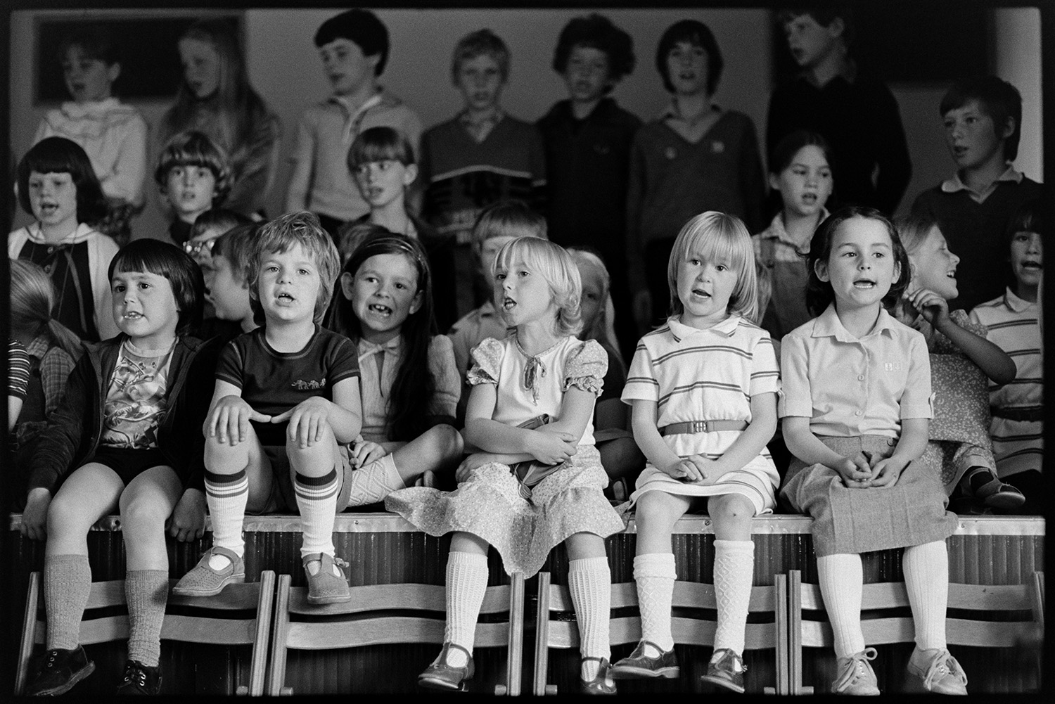 Schoolchildren giving concert in village hall, headmistress and children singing. 
[Schoolchildren singing in a concert or event in Dolton Village Hall. They are sitting on the stage in the hall.]