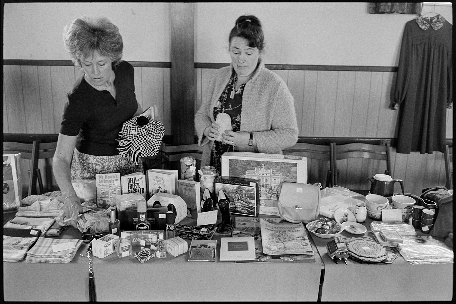 Schoolchildren giving concert in village hall, headmistress and children singing. 
[Two women running a bric-a-brac stall in an event at Dolton Village Hall. Various items are visible including books, a jigsaw and bags.]
