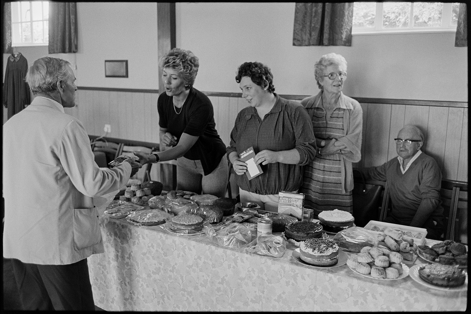 Schoolchildren giving concert in village hall, headmistress and children singing. 
[A man buying a cake from two women running a cake stall in an event at Dolton Village Hall.]