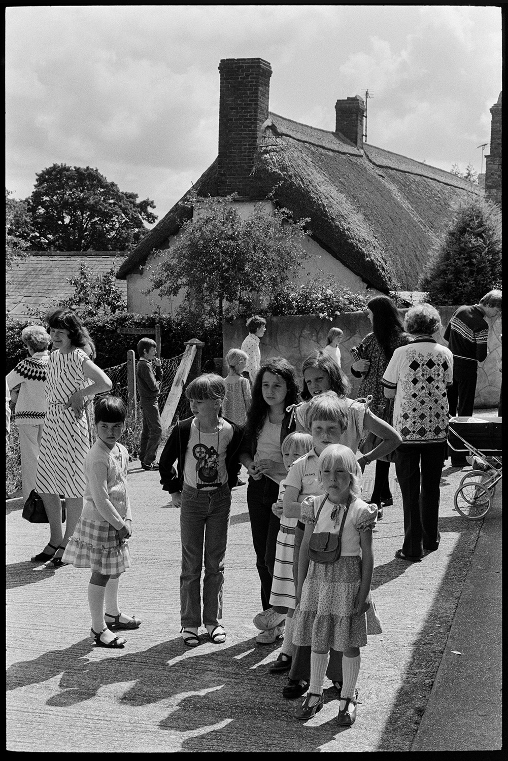 Children waiting for pony ride outside village hall. 
[Children lined up waiting for a pony ride at Dolton Village Hall. Women are waiting nearby and a thatched cottage can be seen in the background.]