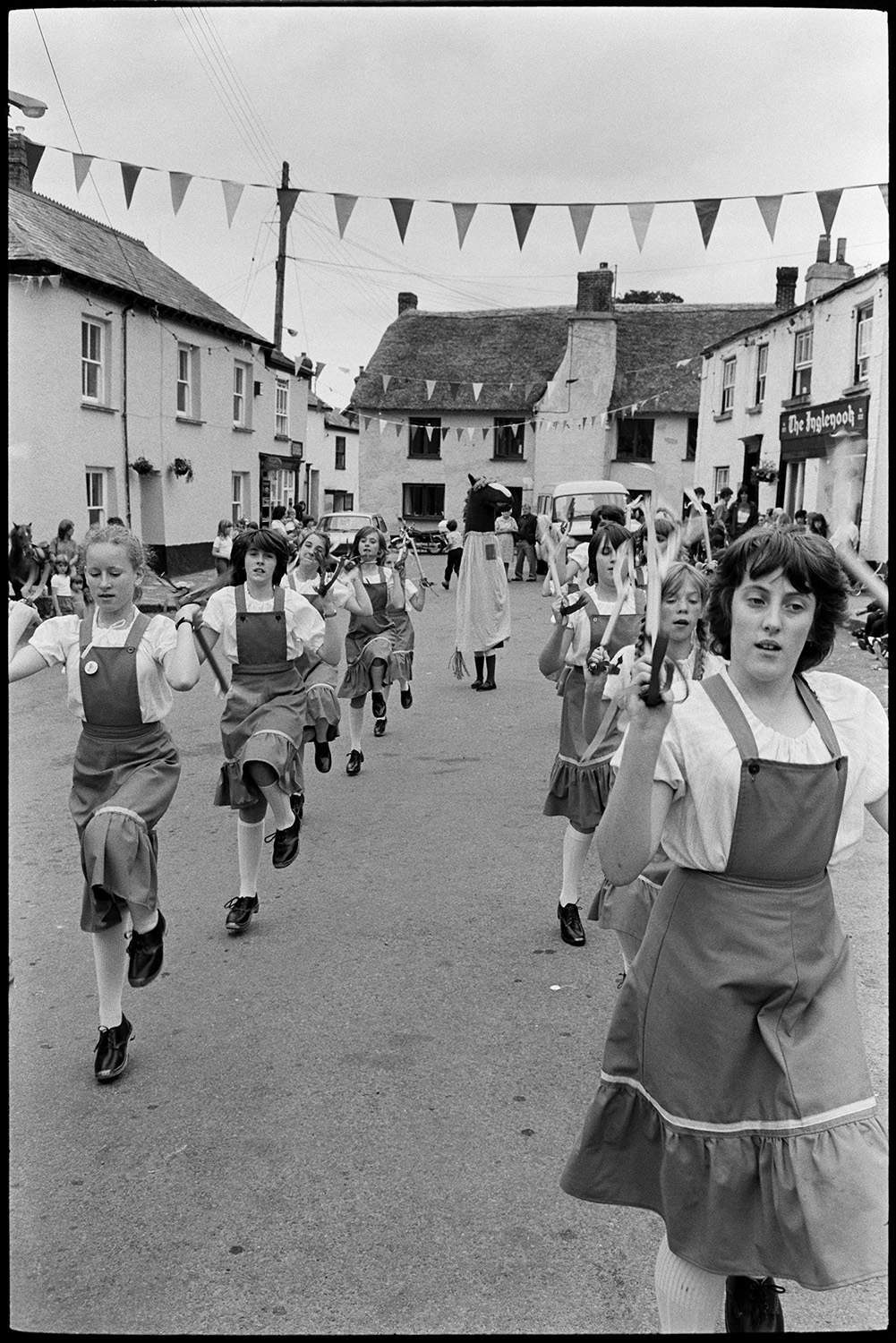 People waiting for fair, cat, pavement artists, Morris Dance.
[A group of girl Morris Dancers or Clog Dancers performing at Winkleigh Fair. A person wearing a horse's head costume is with them. Bunting is strung across the street and  cottages are visible in the background, as well as 'The Inglenook'.]