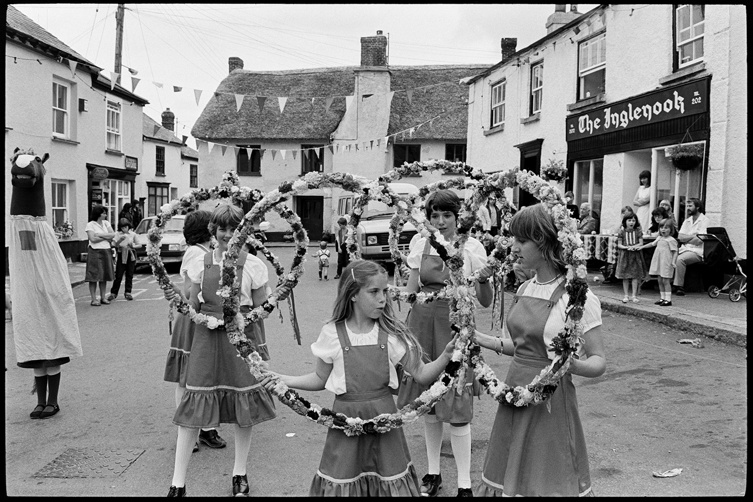 People waiting for fair, cat, pavement artists, Morris Dance.
[A group of girl Morris Dancers or Clog Dancers performing with floral hoops during Winkleigh Fair. A person wearing a horse's head costume is with them. Spectators are watching them from the roadside. A thatched cottage, post office, and the 'The Inglenook' are visible in the background.]