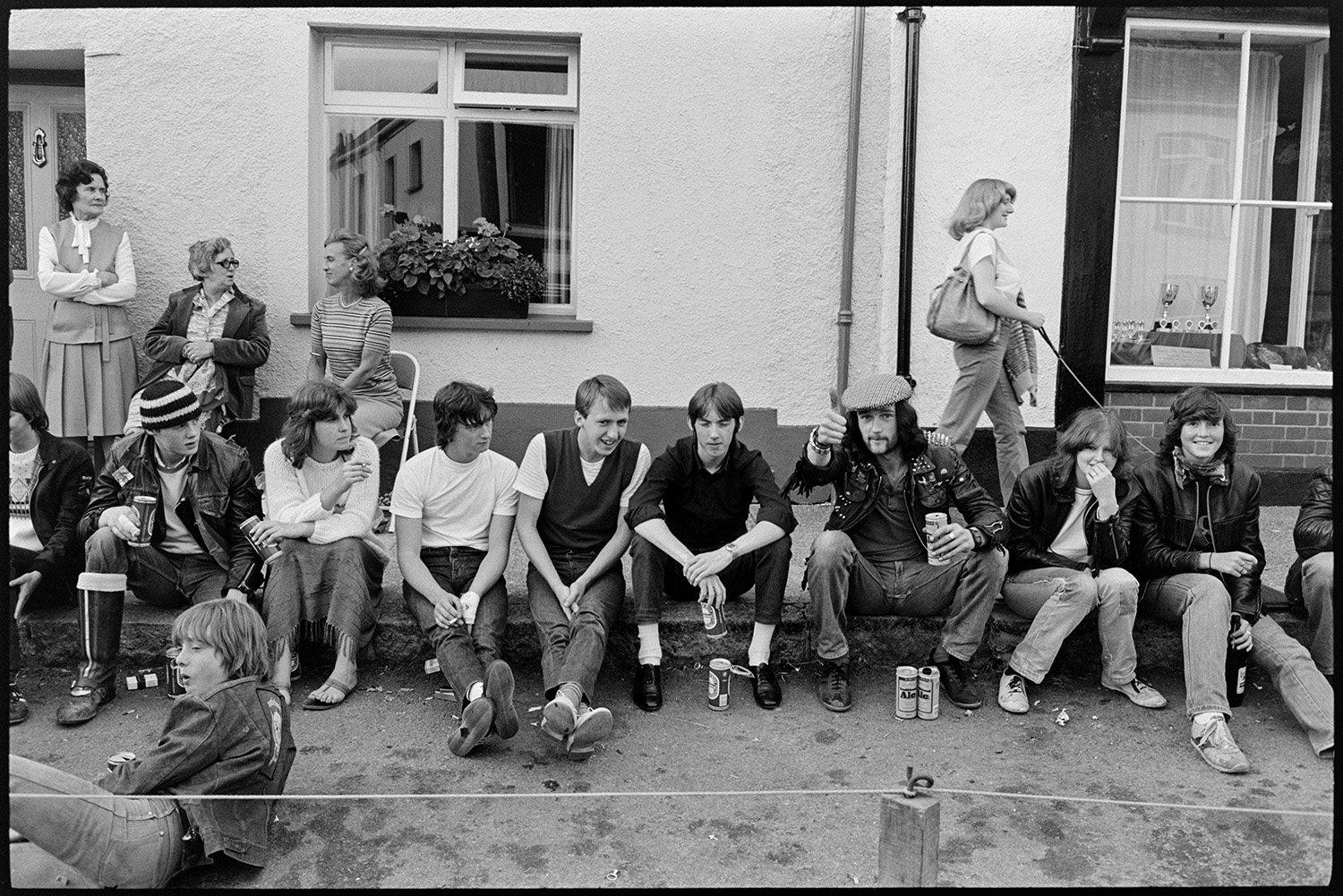 People waiting for fair, cat, pavement artists, Morris Dance.
[Young men and women sitting in a row on a pavement during Winkleigh Fair.  Some are drinking cans of beer and wearing leather jackets. Three ladies are talking behind them, and a woman is walking her dog. A window behind them shows a display of trophies, and another has a window box of plants.]