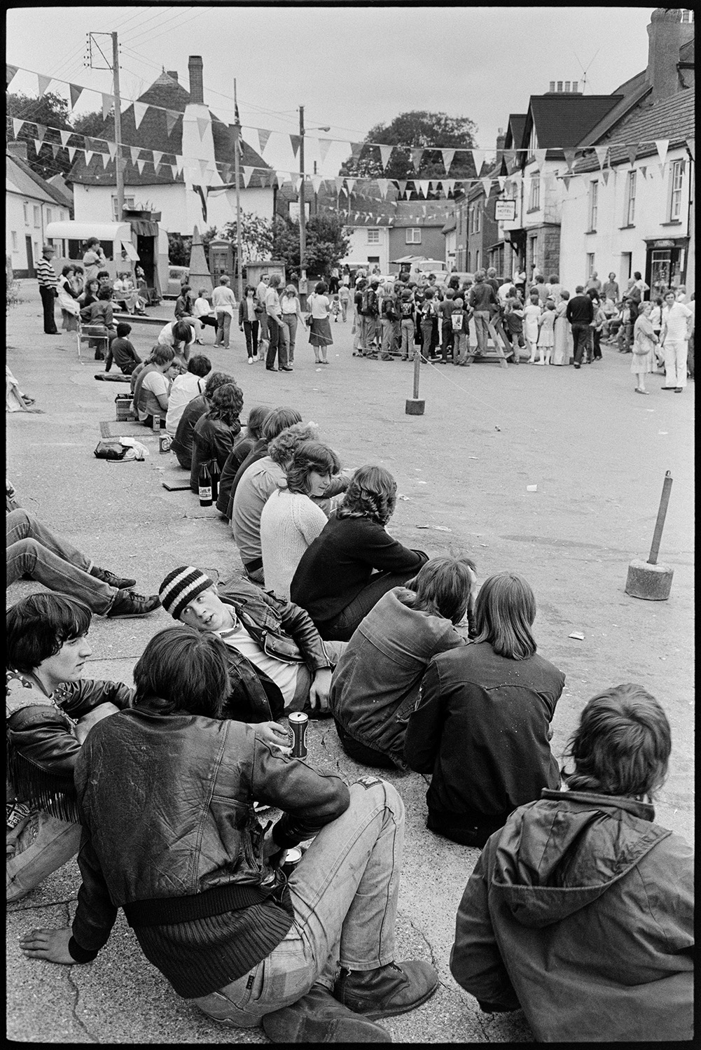 People waiting for fair, cat, pavement artists, Morris Dance.
[Young people, including teenagers in leather jackets, sitting on a pavement in Winkleigh Square, waiting for Winkleigh Fair to start. Bunting is strung across the square, and a crowd of people gathered further along the street.]