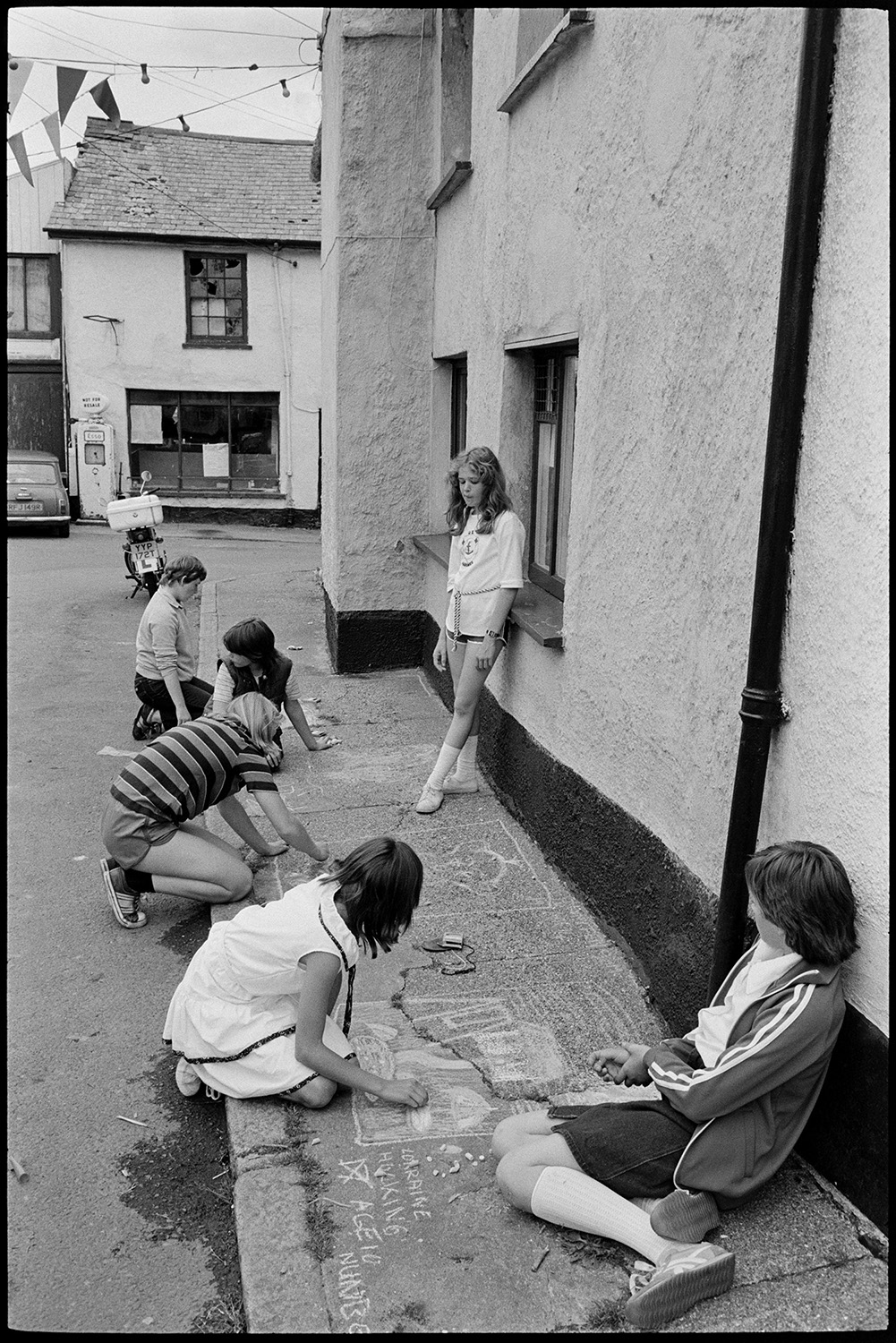 People waiting for fair, cat, pavement artists, Morris Dance.
[Six children, drawing on a pavement with chalk during Winkleigh Fair. A mini car, moped, petrol pump, bunting and cottages are visible in the background.]