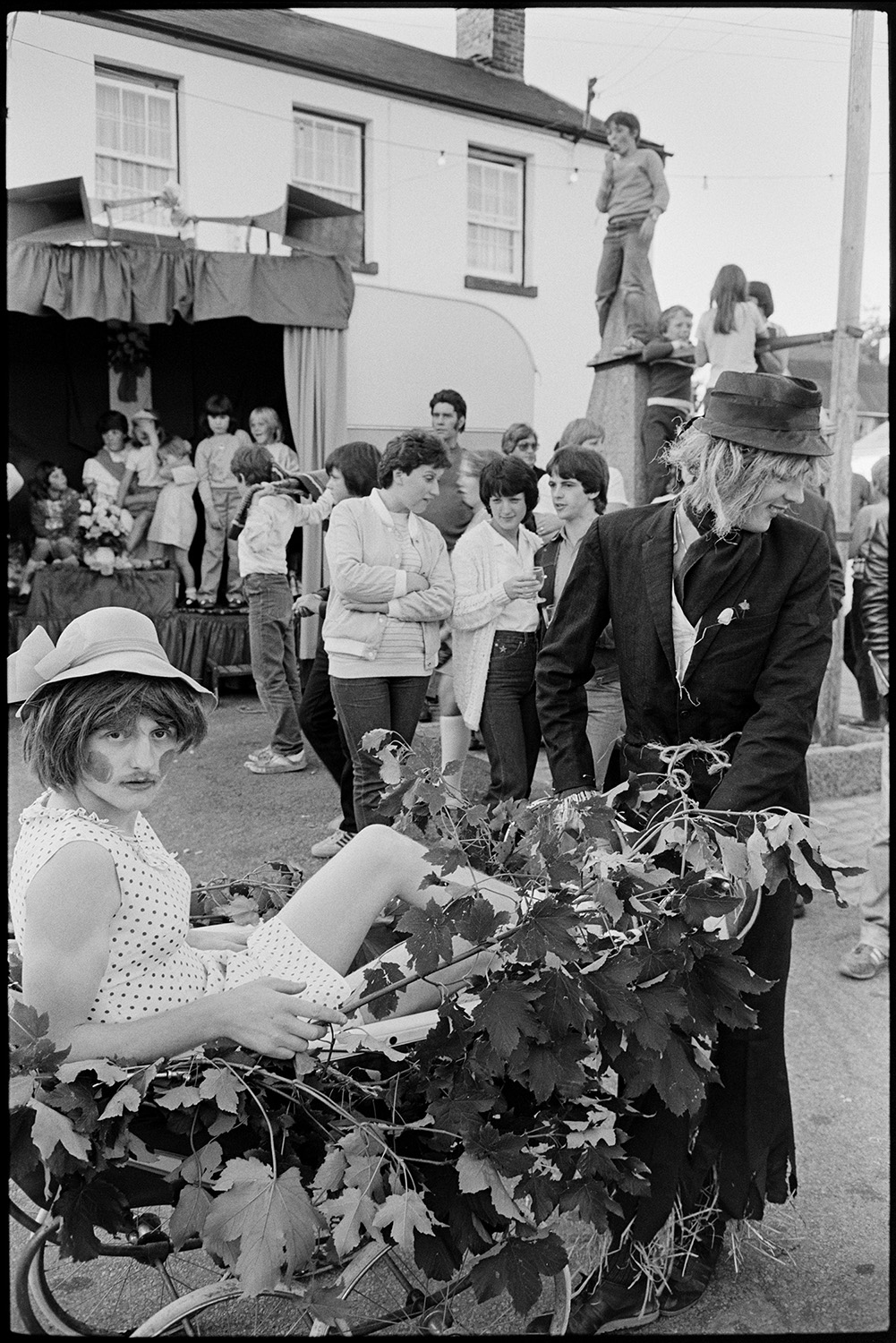 Fancy dress, pram race around village.
[Contestants in fancy dress waiting for the start of the pram race at Winkleigh Fair. One entry has an old pram covered in leaves. People are talking and watching in the background, by the podium for the Fair Queen and a boy stood on a stone pillar to get a better view.]