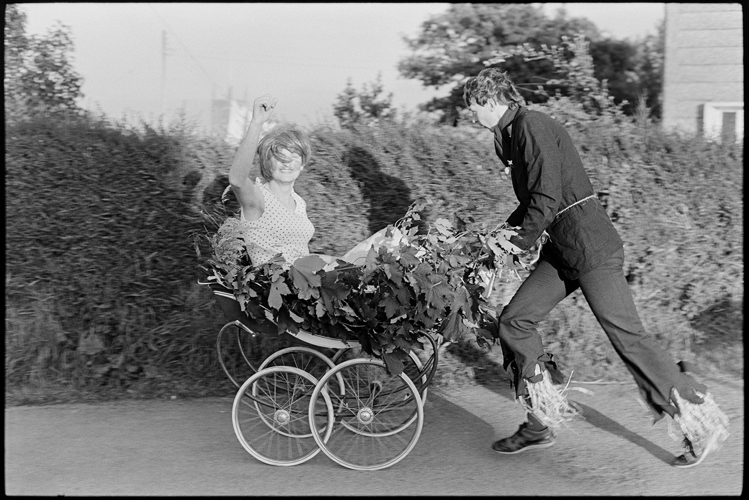 Fancy dress, pram race around village.
[Two people in fancy dress competing in a pram race around the village during Winkleigh Fair. One person is riding in the old pram covered in leaves, while the other pushes it.]