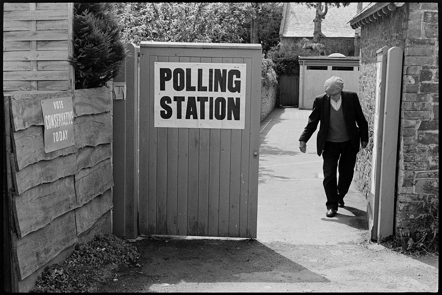 Notice outside polling station on election day.
[Man walking out of Dolton School polling station on election day. A 'Polling Station' notice is pinned to a  wooden gate, and a voting poster is stuck to a wooden fence.]