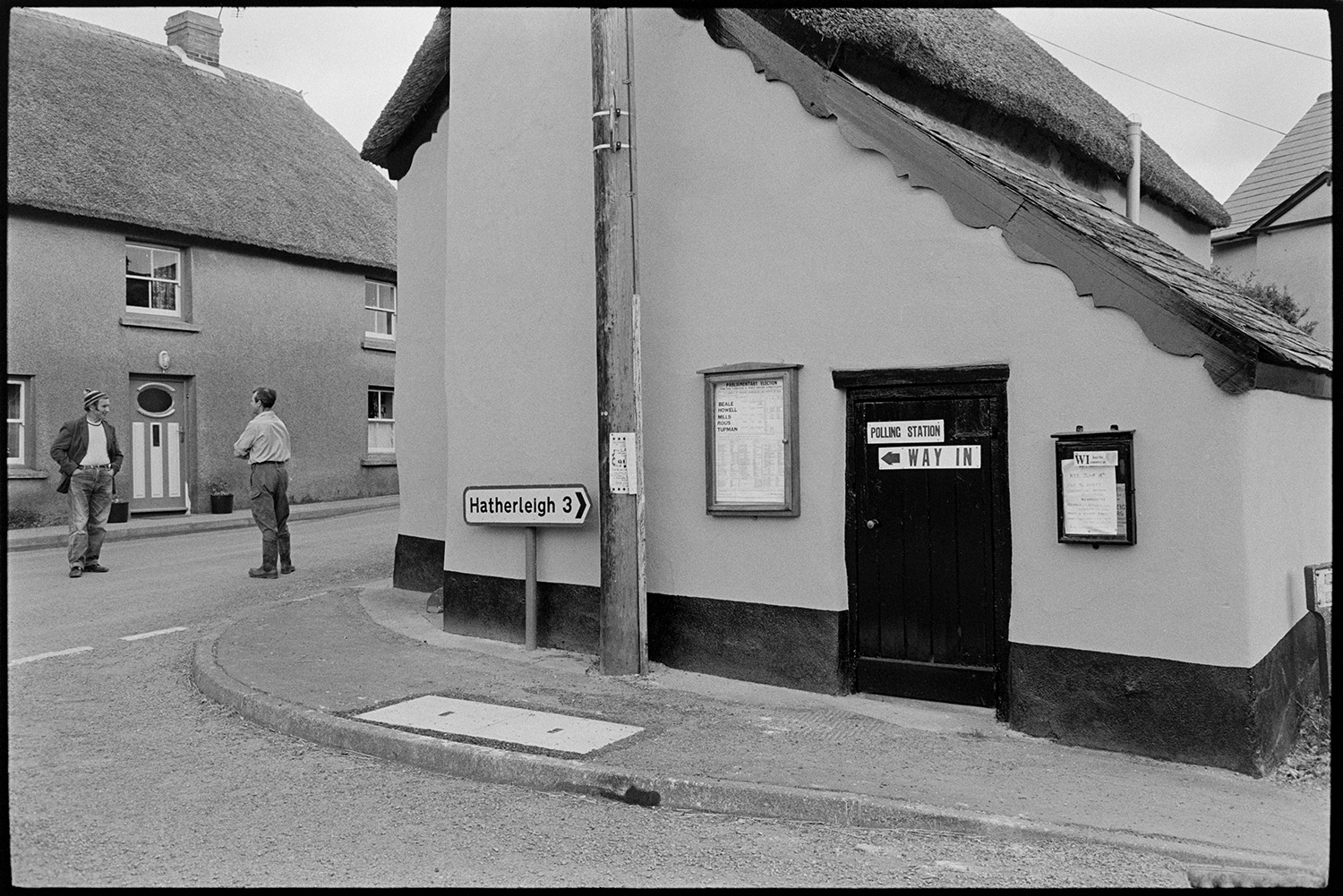 Notices on village polling station.
[A thatched cottage with a 'Polling Station' poster and notice boards attached to its wall, at Monkokehampton. Two men are talking in street outside.]