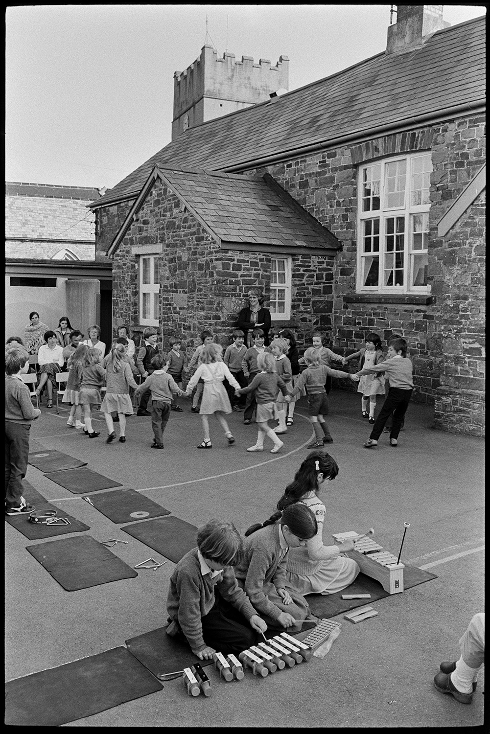 Schoolchildren dancing in school playground. 
[Schoolchildren playing xylophones and dancing in a circle in Dolton Primary School playground. The school building and church tower can be seen in the background.]