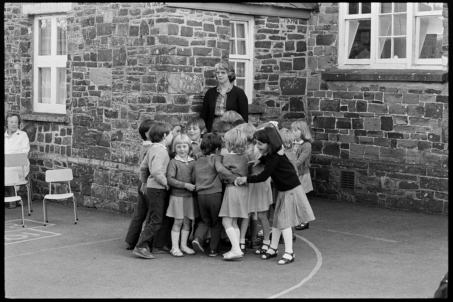 Schoolchildren dancing in school playground. 
[A woman supervising schoolchildren playing in Dolton Primary School playground. She is leaning against part of the school building.]