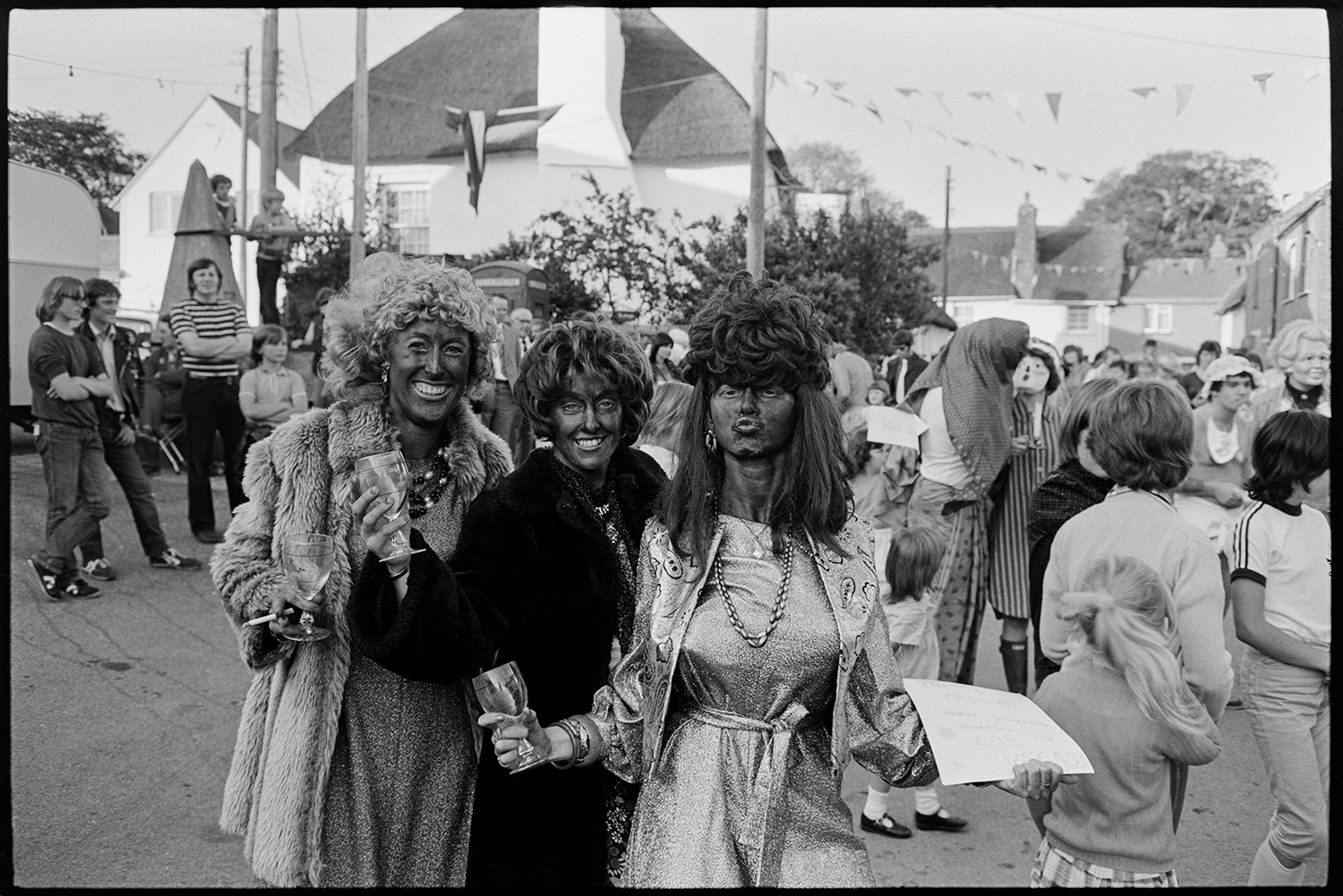 Village Fair, folk clog dancing. Fancy dress, spectators. 
[Three women in fancy dress and holding wine glasses at Winkleigh Fair. Other people, some also in fancy dress, can be seen in the background. Winkleigh square is decorated with bunting.]