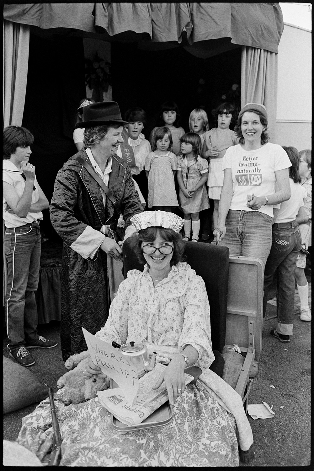 Village Fair, folk clog dancing. Fancy dress, spectators. 
[A woman in fancy dress as Queen Elizabeth II at Winkleigh Fair. She is holding a sign reading 'Where's Philip?'. A group of children are gathered on a stage in the background.]