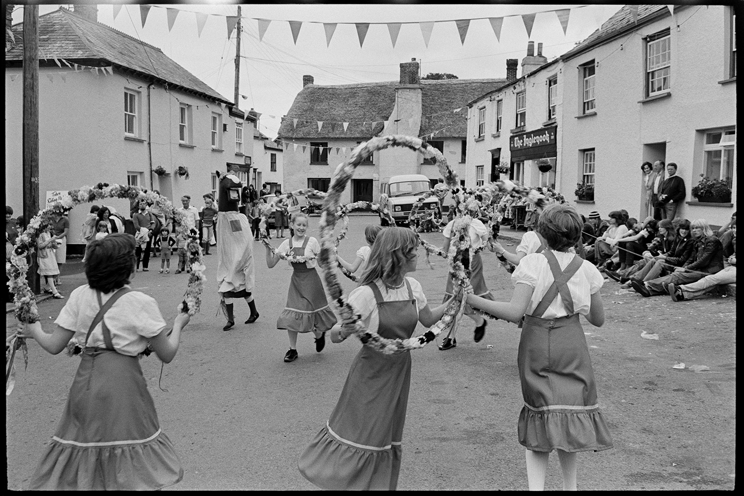 Village Fair, folk clog dancing. Fancy dress, spectators. 
[A group of girl Morris Dancers or Clog Dancers performing with floral garlands at Winkleigh Fair. People are gathered around Winkleigh Square, which is decorated with bunting, watching them. A person wearing a horse's head costume is with them.]