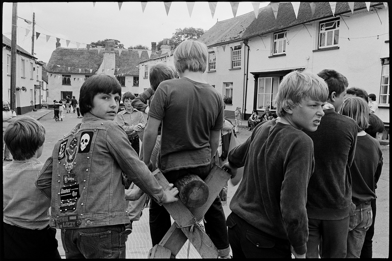 Village Fair, folk clog dancing. Fancy dress, spectators. 
[Teenage boys playing on a greasy pole game at Winkleigh Fair. One boy is wearing a jacket with badges and the street is decorated with bunting.]