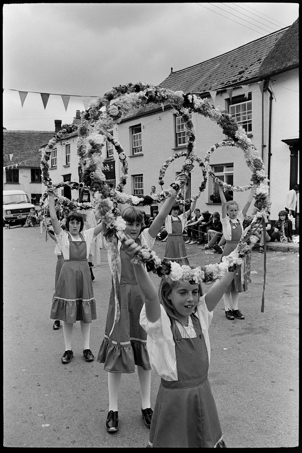 Village Fair, folk clog dancing. Fancy dress, spectators. 
[A group of girl Morris Dancers or Clog Dancers performing with floral garlands at Winkleigh Fair. People are watching them in Winkleigh Square and the street is decorated with bunting.]