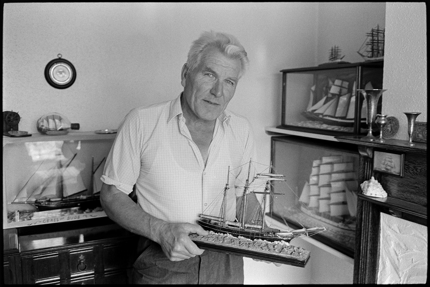 Retired sailor and wife, posing and workshop with model ships. 
[A retired sailor holding a model ship which he has made, in his house at Braunton. Other model ships are displayed in glass cases behind him.]