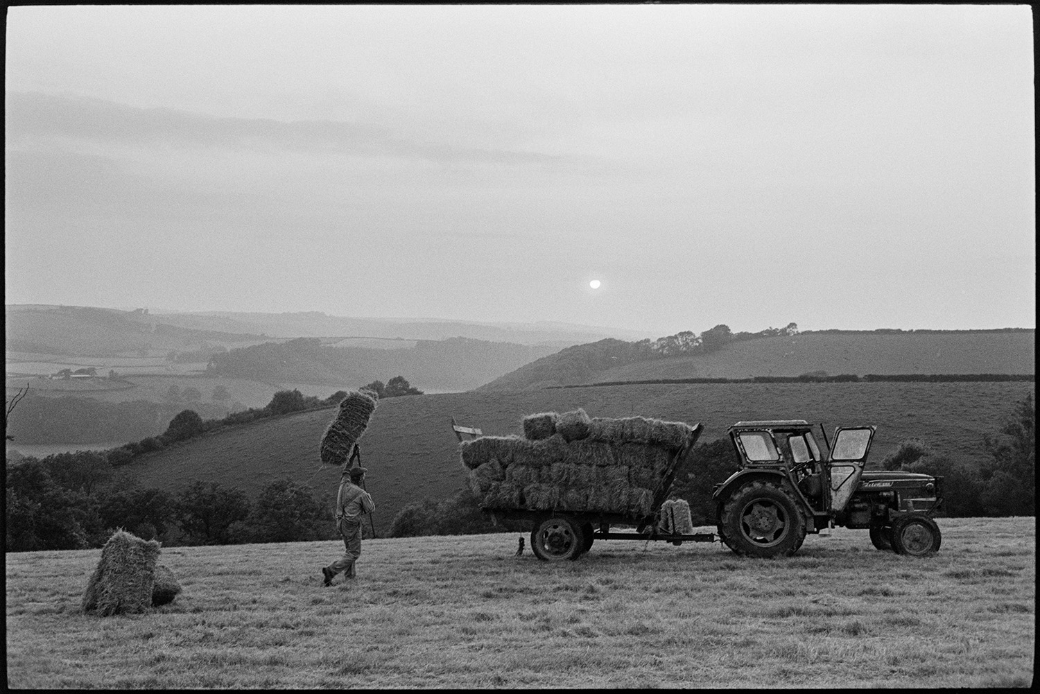 Farmer loading hay bales onto trailer, evening. 
[George Ayre loading hay bales onto a tractor and trailer in a field at Ashwell, Dolton, in the evening. He is carrying a hay bale above his head using a pitchfork. Fields can be seen on a hillside in the background.]