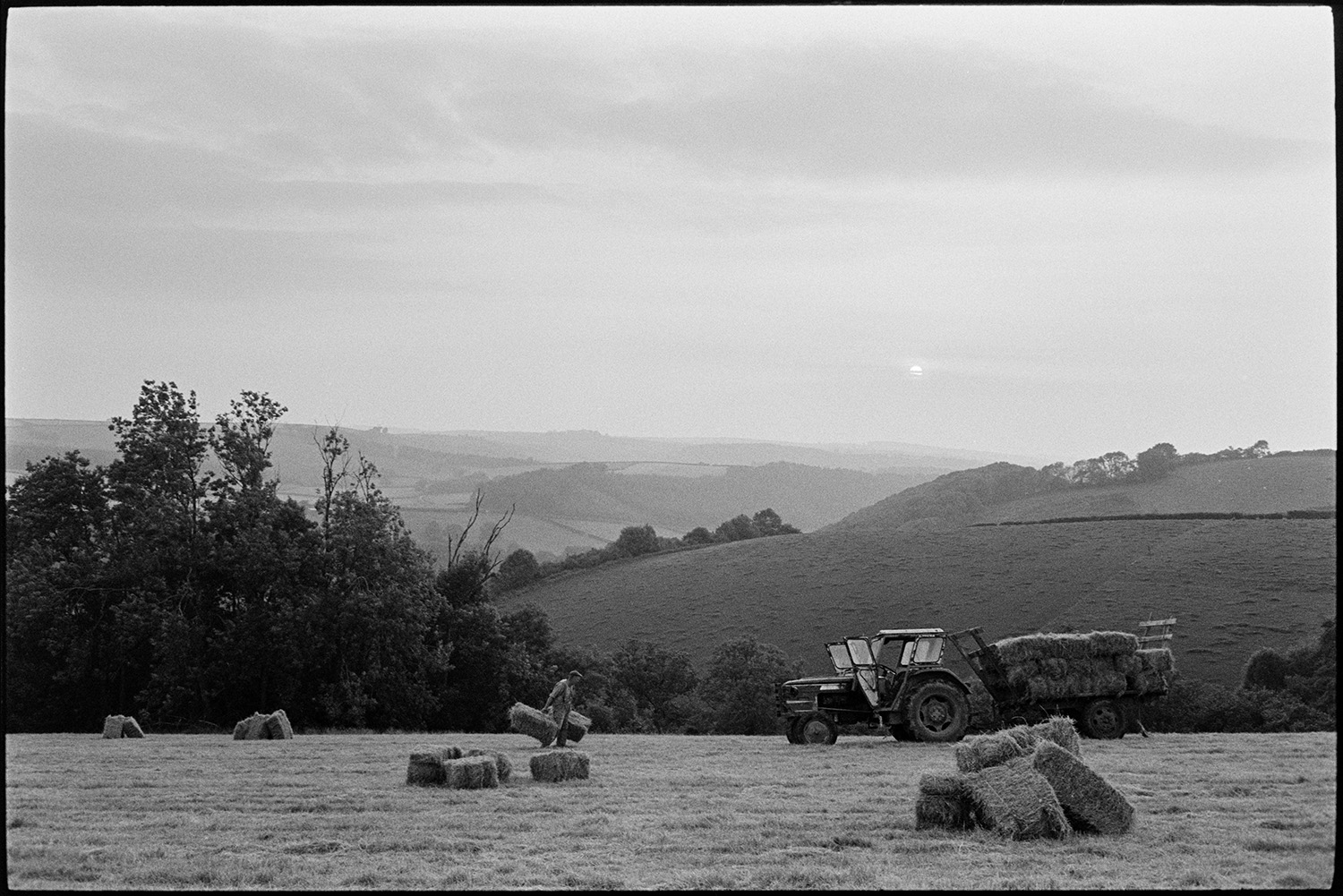 Farmer loading hay bales onto trailer, evening. 
[George Ayre loading hay bales onto a tractor and trailer in a field at Ashwell, Dolton, in the evening. Stacks of hay bales can be seen in the field and fields are visible on a hillside in the background.]