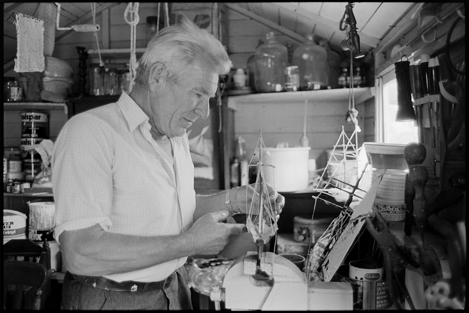 Retired sailor and wife, posing and workshop with model ships. 
[A retired sailor working on a model ship in his workshop at Braunton.  Parts of models, paint tins and glass jars can be seen in the background.]