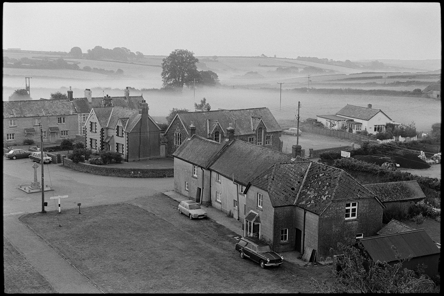 View of village from church tower, early mist. 
[A view of houses and parked cars in Ashreigney taken from Ashreigney Church tower in the early morning. The war memorial can be see in the village square and misty fields are visible in the background.]