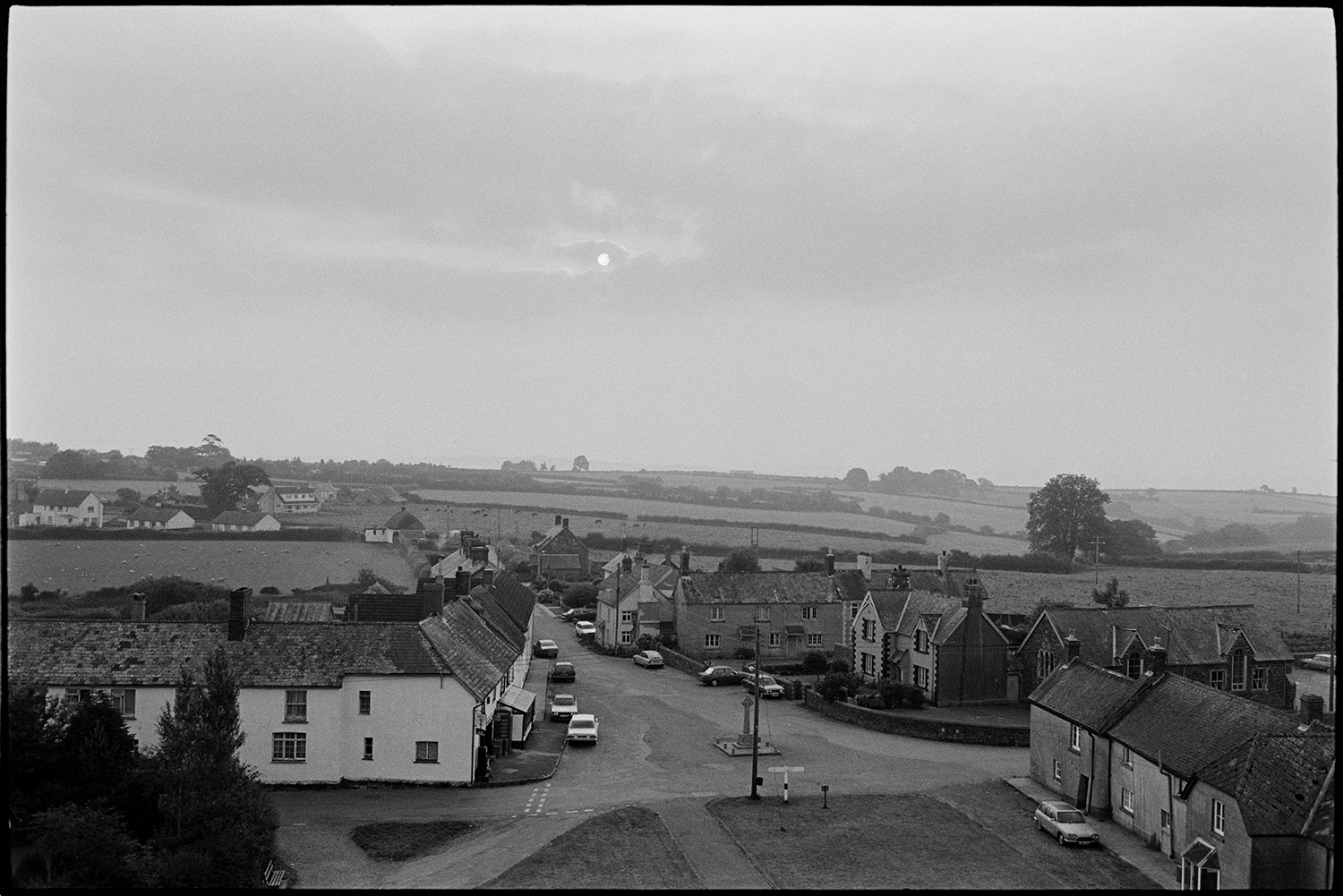 View of village from church tower, early mist. 
[A view of houses and parked cars in Ashreigney taken from Ashreigney Church tower in the early morning. The war memorial can be see in the village square and misty fields with grazing livestock are visible in the background.]