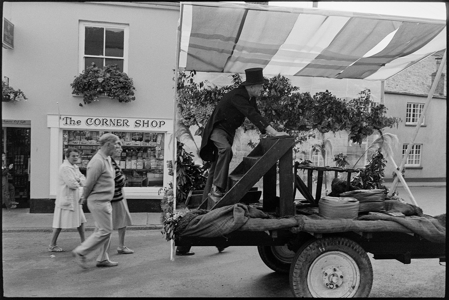 Village Fair, start of race for children in street, women, mothers holding children. 
[A man wearing a top hat on a decorated trailer at Chulmleigh Fair. Three people are walking behind the trailer past the shop front of 'The Corner Shop'.]