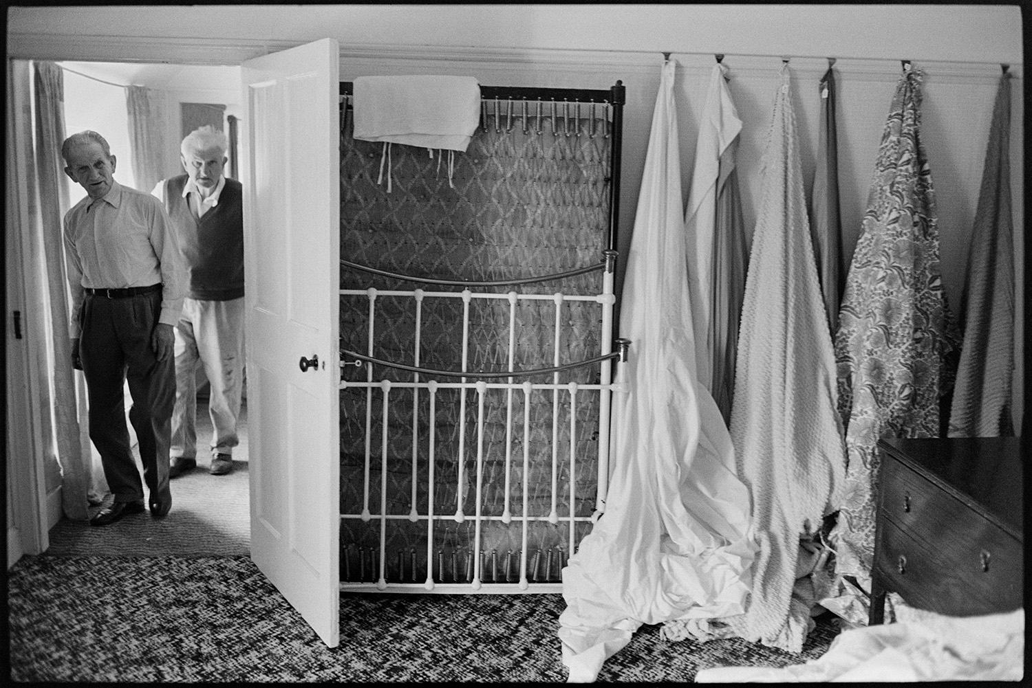 People viewing house contents before sale, bedroom furniture, lace, material. 
[Two men looking at items in Arscotts House, Dolton, on the viewing day before the contents sale of the house. A mattress, bed frame and fabric is displayed against one of the bedroom walls.]