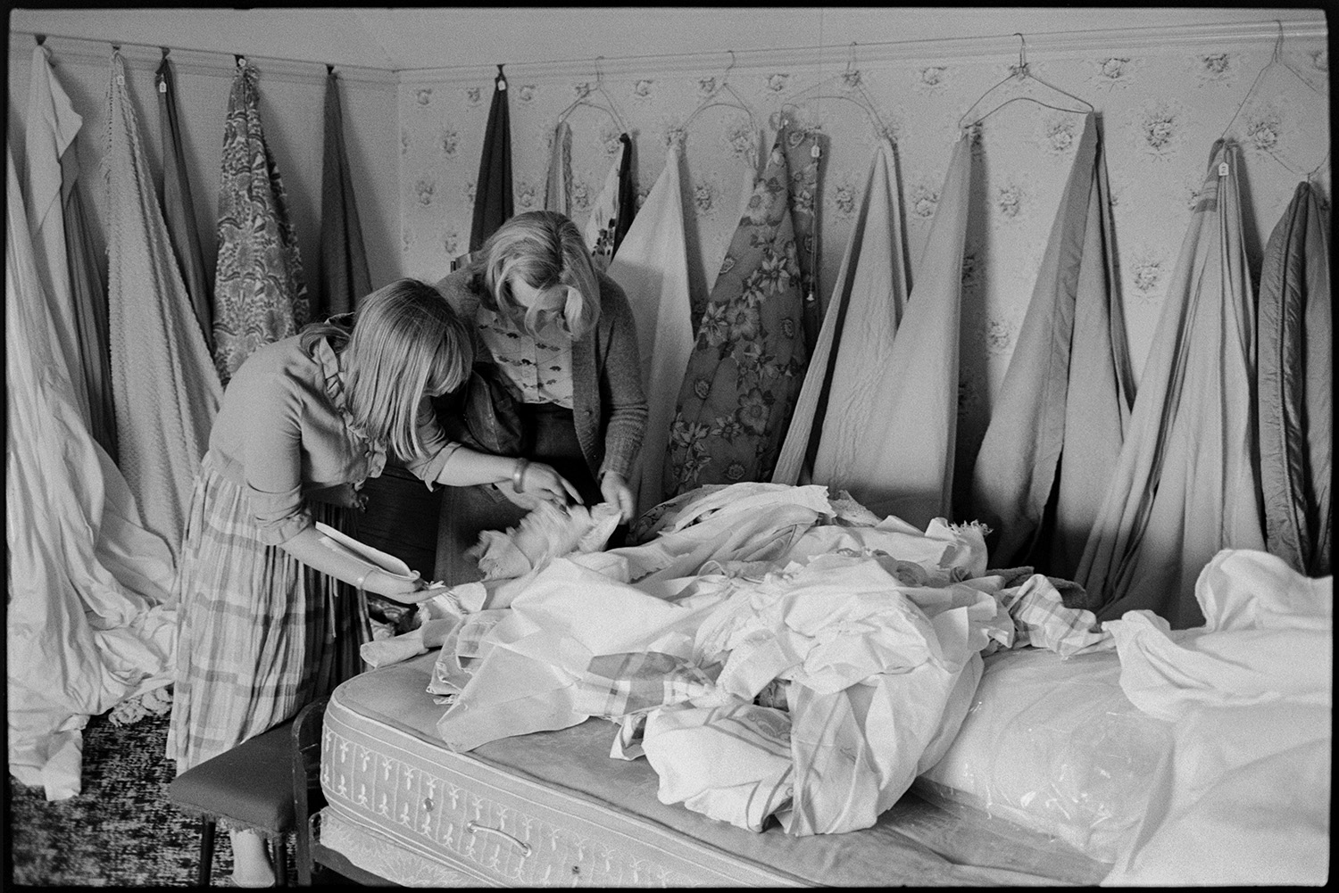 People viewing house contents before sale, bedroom furniture, lace, material. 
[Two women looking through fabric on a mattress at Arscotts House, Dolton, on the viewing day before the contents sale of the house. Material is also hung up on the walls of the bedroom.]