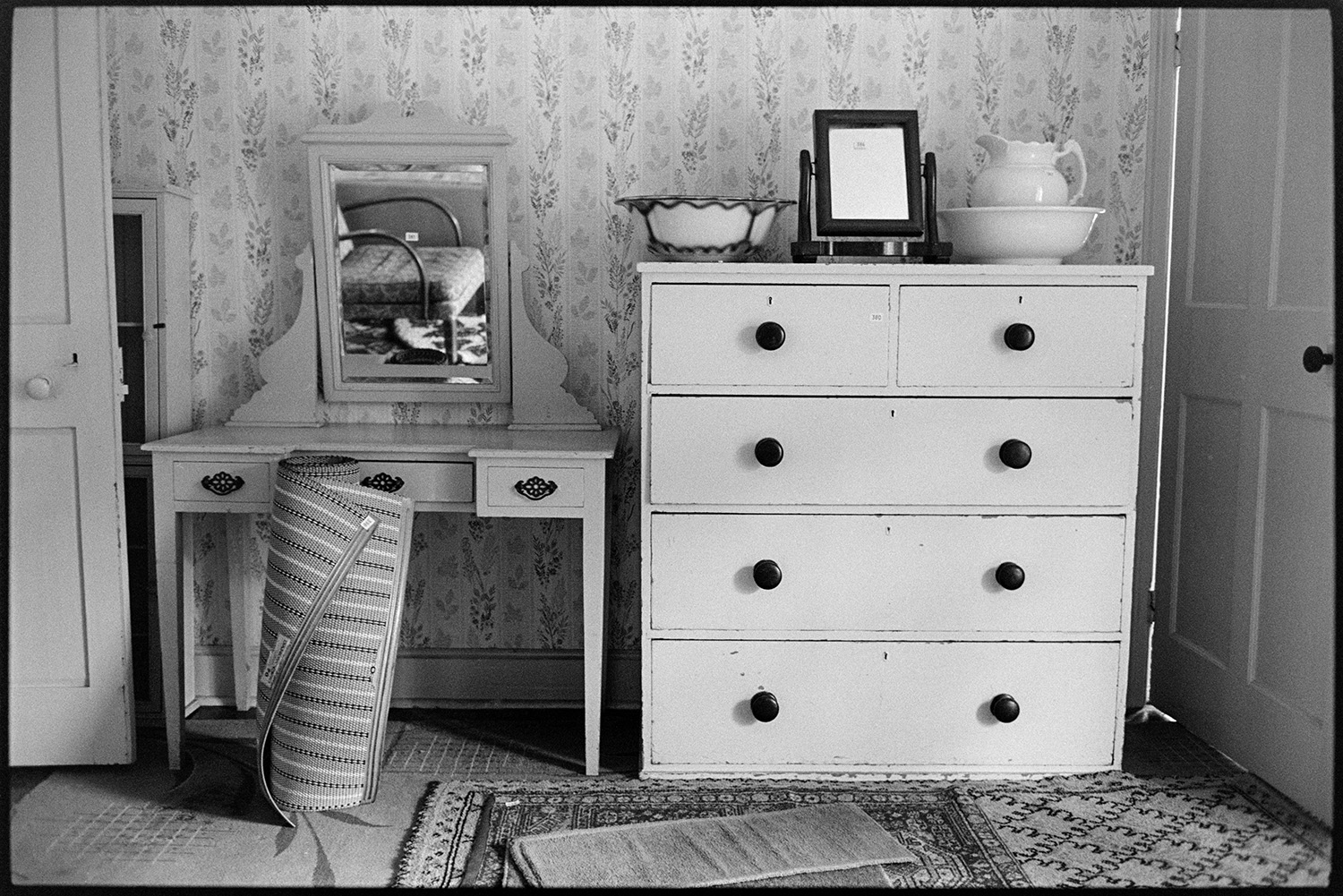 People viewing house contents before sale, bedroom furniture. Pot under bed, women and lace. 
[Items in Arscotts House, Dolton on the viewing day before the contents of the house are sold. A dressing table, chest of drawers, mirror, chamber pot and rugs are visible. The wall is covered with patterned wallpaper.]