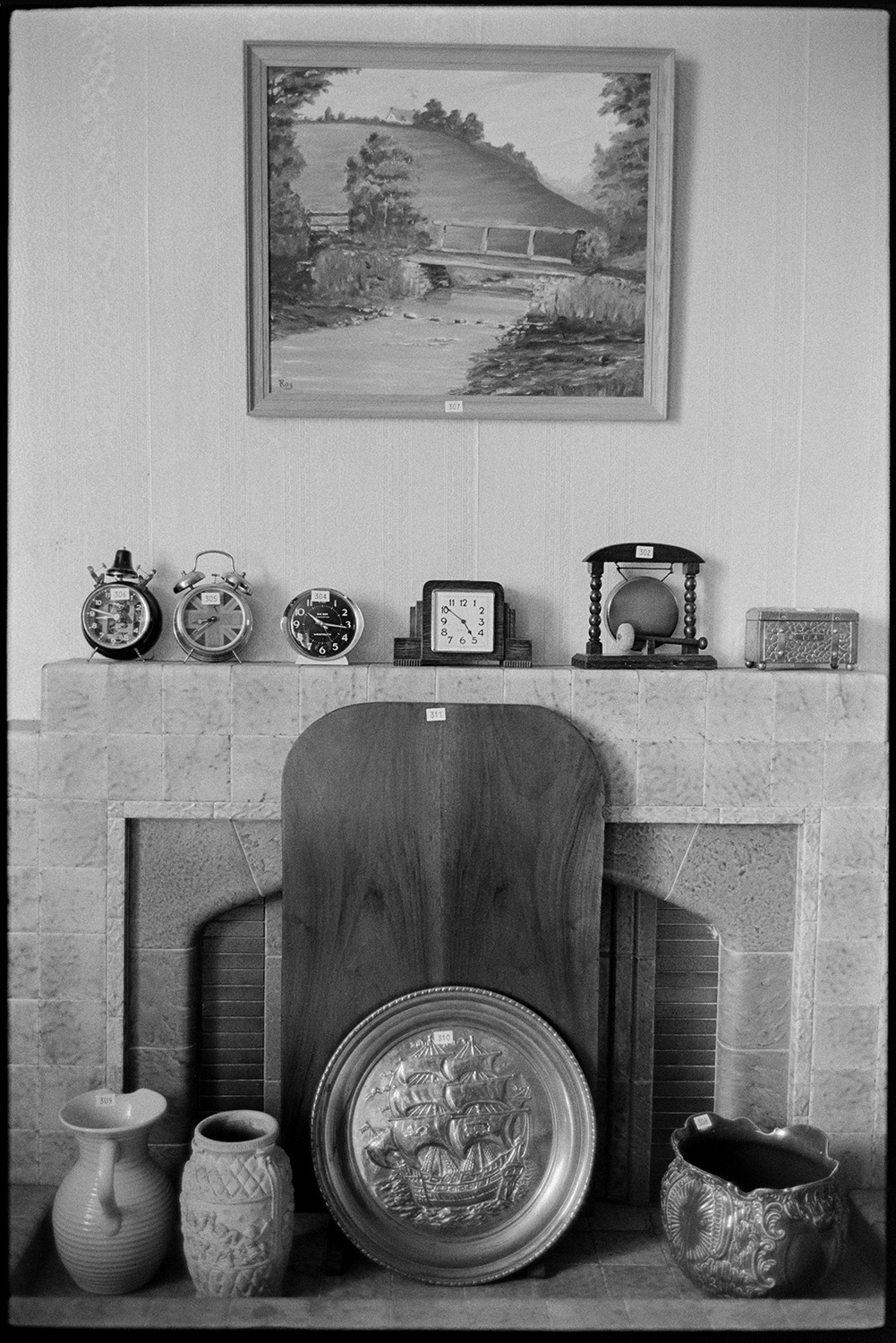 China, clocks and ornaments on viewing day, before house sale fireplace, painting. 
[Jugs, clocks, a gong, metal platter with a ship design and a painting displayed by a fireplace at Arscotts House, Dolton, on the viewing day before the contents of the house are sold.]