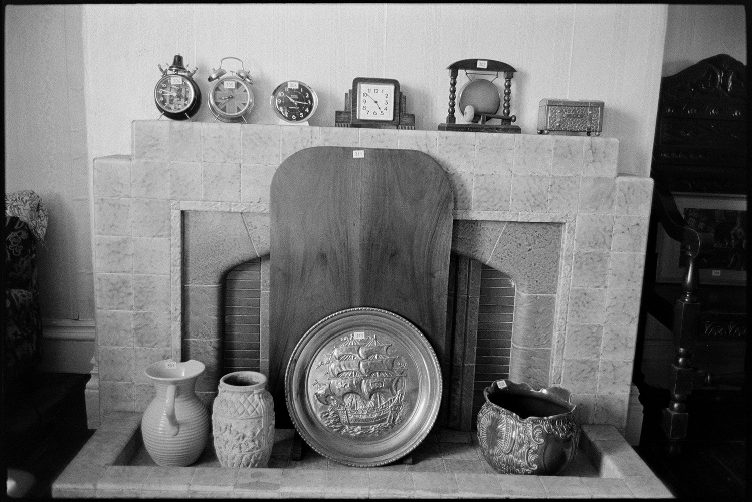 China, clocks and ornaments on viewing day, before house sale fireplace, painting. 
[ A close up view of jugs, clocks, a gong and a metal platter with a ship design displayed by a fireplace at Arscotts House, Dolton, on the viewing day before the contents of the house are sold.]