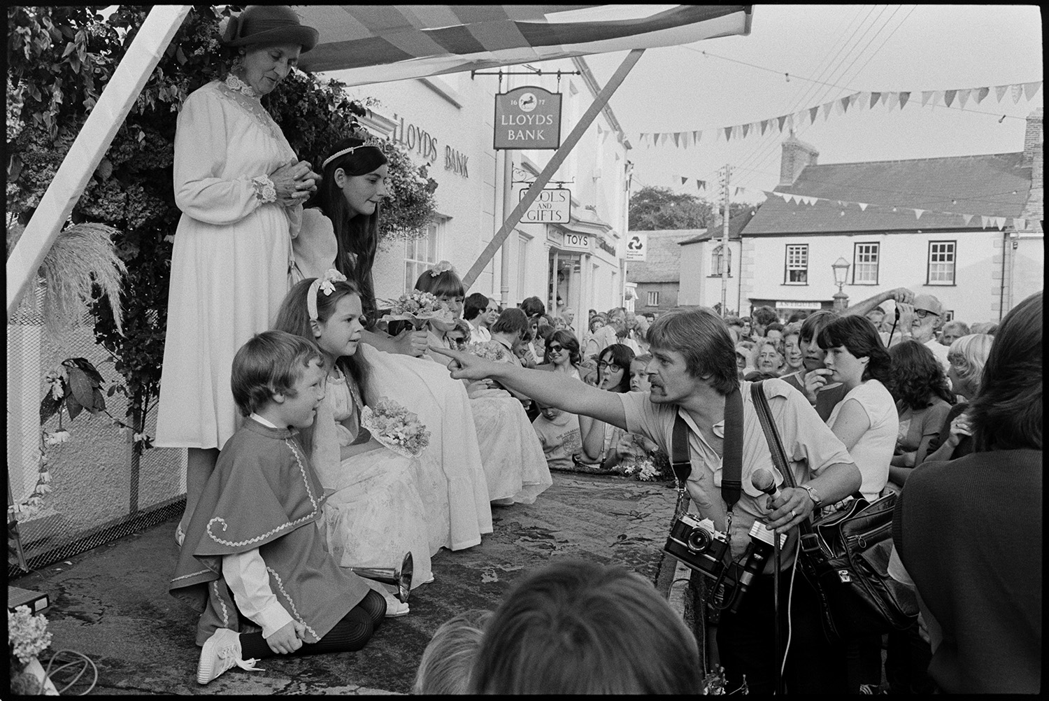 Procession with Fair Queen and band, speech from window, raising the glove, spectators. 
[A man arranging the Chulmleigh Fair Queen and her attendants sat on a podium outside Lloyds Bank, with another woman, at Chulmleigh Fair. He is going to photograph or film them. People are gathered around watching.]
