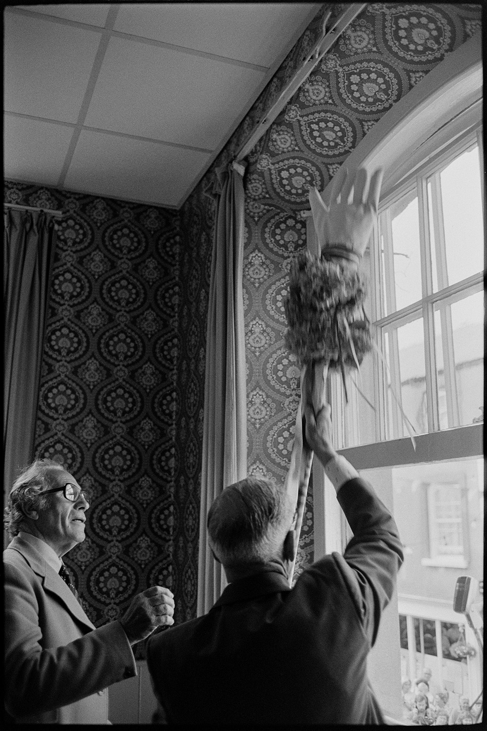 Procession with Fair Queen and band, speech from window, raising the glove, spectators. 
[Joe Fullbrook stood next to another man lifting the Chulmleigh Fair glove by a window inside the town hall, at Chulmleigh Fair. The walls of the room are covered with patterned wallpaper.]