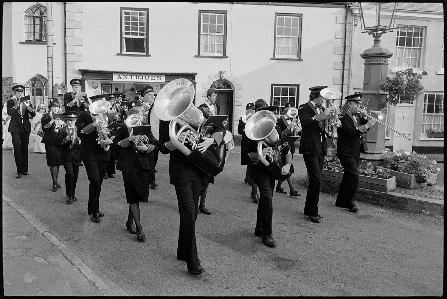 Procession with Fair Queen and band, speech from window, raising the glove, spectators. 
[A brass band marching along a street in Chulmleigh at Chulmleigh Fair. They are passing an Antiques Shop window and a lamp decorated with flowers in planters.]