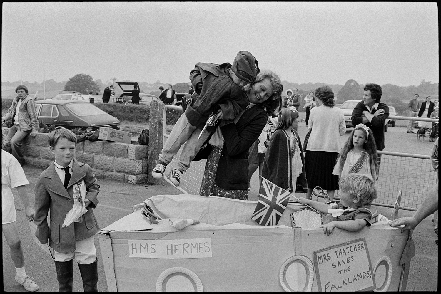 Fancy dress preparations, queen and attendants preparing for parade with brass band. 
[A woman placing a child into a fancy dress entry of a pram made into cardboard boat of HMS Hermes, at Chulmleigh Fair. A sign on the side reads 'Mrs Thatched Saves the Falklands!'. Other people and parked cars can be seen in the background.]