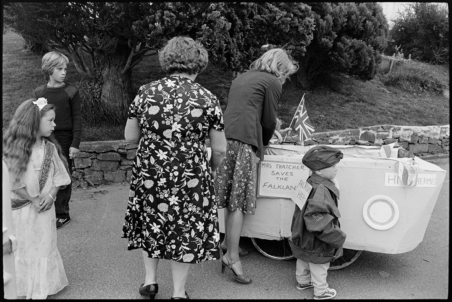Fancy dress preparations, queen and attendants preparing for parade with brass band. 
[A woman placing a child into a fancy dress entry of a pram made into cardboard boat of HMS Hermes, at Chulmleigh Fair. A sign on the side reads 'Mrs Thatched Saves the Falklands!' and a child is stood next to her with another sign reading 'Falklands Hero!'. The Fair Princess is watching on.]