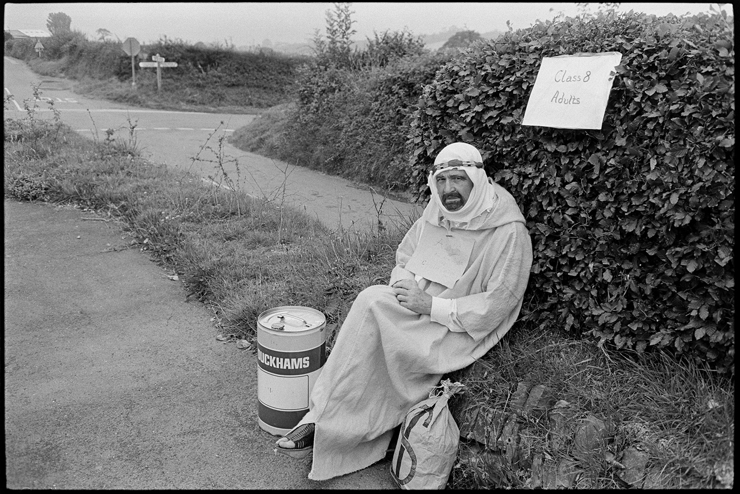 Fancy dress preparations, queen and attendants preparing for parade with brass band. 
[A man in fancy dress as an oil sheik wearing a robe or thawb. He is sat by a roadside with a round tin and a sack with a dollar sign drawn onto it, at Chulmleigh Fair.]