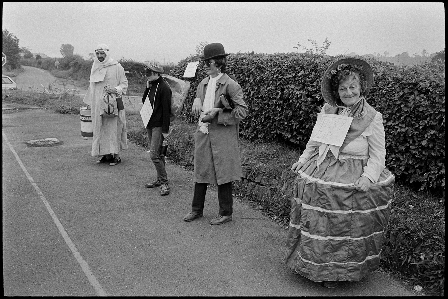 Fancy dress preparations, queen and attendants preparing for parade with brass band. 
[Four adults in fancy dress at Chulmleigh Fair. One man is dressed as an oil sheik and another as a thief or robber.]