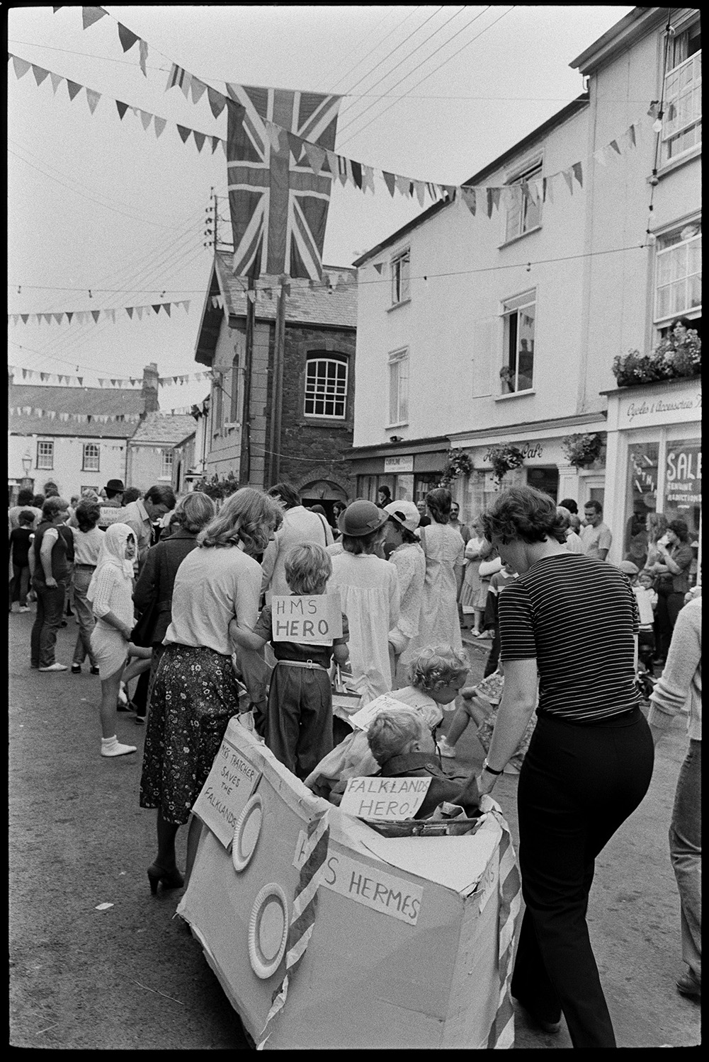 Fancy dress parade with brass band, going home afterwards. 
[Two women pushing a pram made into the shape of the boat HMS Hermes for a fancy dress competition at Chulmleigh Fair. Three children are sat in the 'boat'. Other people in the street are in fancy dress and the street is decorated with bunting and a Union Jack flag.]]