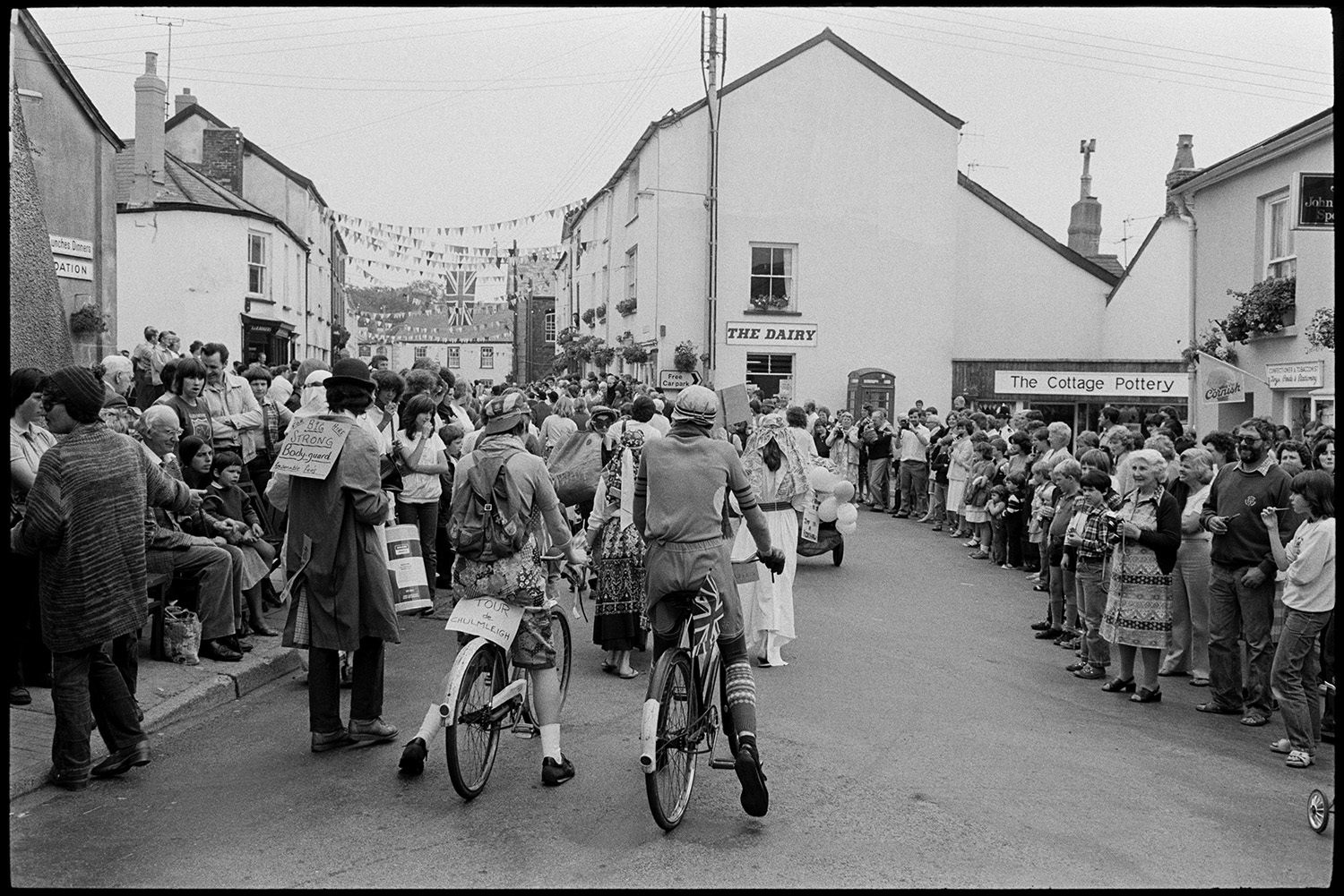 Fancy dress parade with brass band, going home afterwards. 
[Two people riding bicycles in a fancy dress parade through a street at Chulmleigh Fair. Their entry is called Tour de Chulmleigh. People are lining the street watching the parade.]