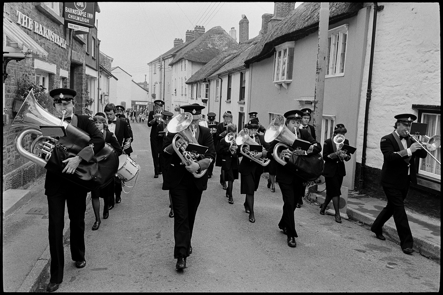 Fancy dress parade with brass band, going home afterwards. 
[A brass band marching along a street at Chulmleigh Fair. They are passing the Barnstaple Inn and thatched cottages.]