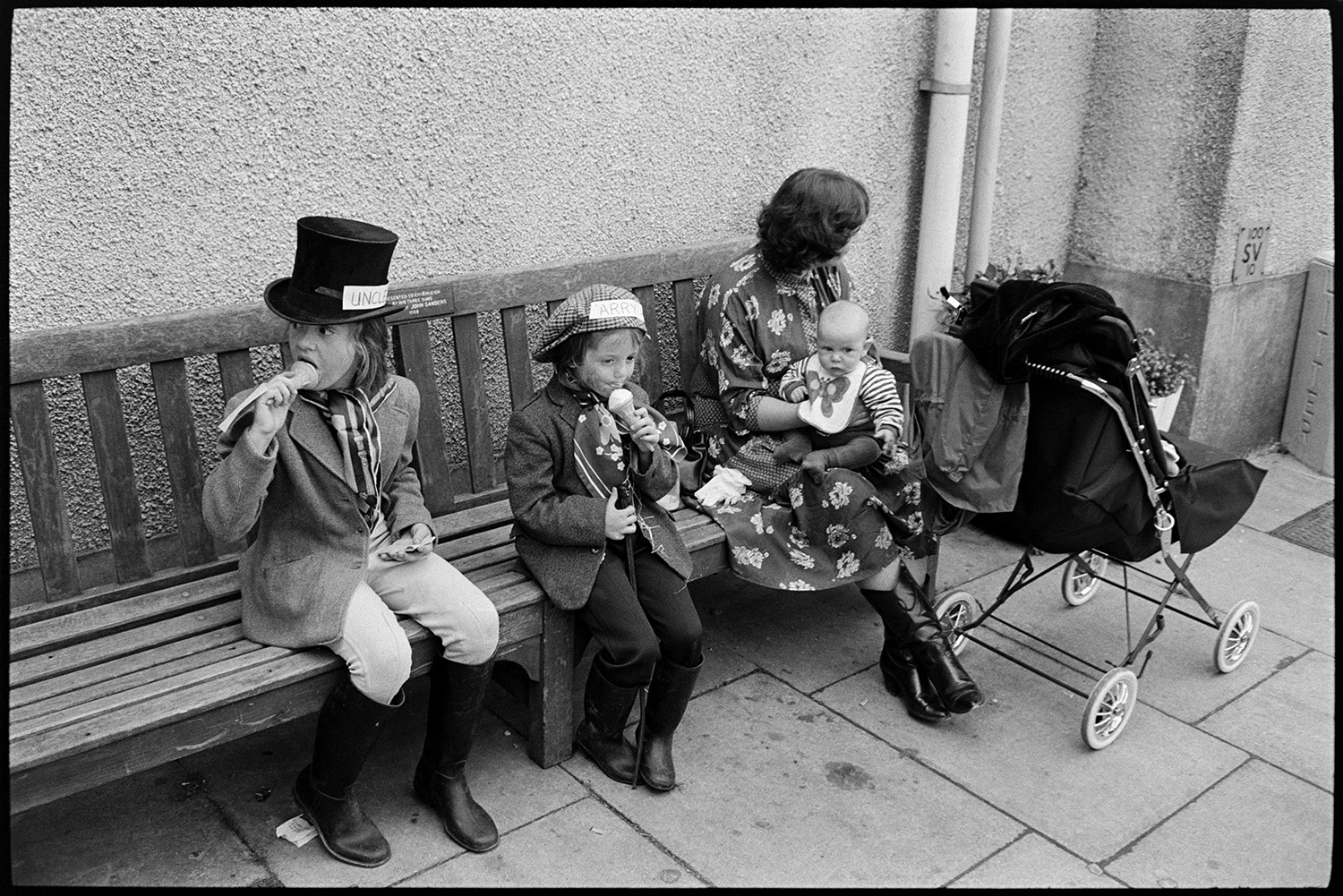 Fancy dress parade with brass band, going home afterwards. 
[A woman with two children in fancy dress, from the 'Widecombe Fair' entry, sat on a bench eating ice creams at Chulmleigh Fair. She is holding a baby on her lap and a pram is parked next o them.]